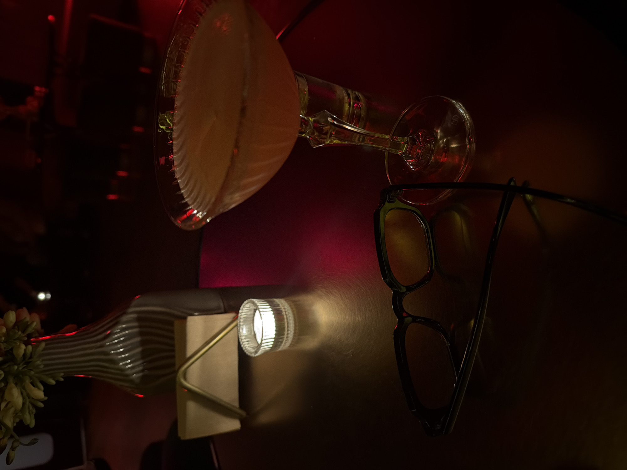 The image captures a dimly lit scene, likely in Yerevan, Armenia, given the user's prompt. A martini glass filled with a light-colored beverage is precariously positioned near a pair of dark-rimmed glasses, suggesting a relaxed or after-hours atmosphere. In the lower portion of the frame, a striped glass vase containing a bouquet of flowers sits next to a small lamp projecting a beam of light. The arrangement creates a juxtaposition of elegance and casualness, possibly in a bar or restaurant setting. The red-tinged lighting adds to the intimate ambiance.