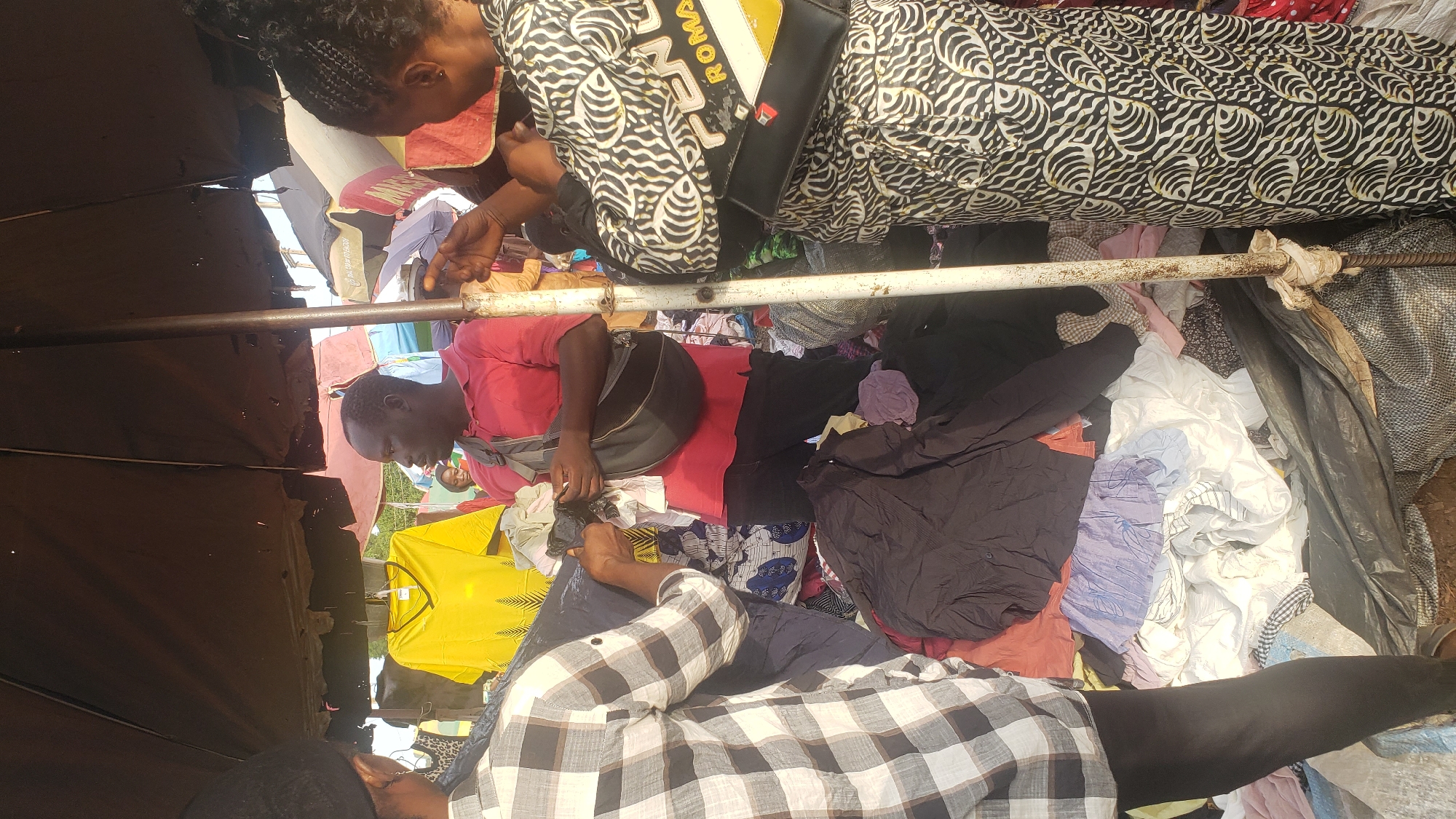 In the city of Kakuri Gwari, Nigeria, a street market scene unfolds under the shade of a large, dark umbrella. The market appears to be an open-air arrangement of goods, primarily clothing, displayed on the ground and on makeshift stalls. Three individuals are prominently visible, suggesting a transaction or interaction is occurring.
A man in a red polo shirt, wearing a dark cross-body bag, stands centrally. To his left, partially obscured, is a person wearing a black and white checkered shirt and a black head covering. This individual is reaching towards a pile of clothing. To the man's right, a woman in a black and white patterned dress with a visible black and yellow bag is facing away from the camera, also seemingly engaged in the market activity.
The foreground is dominated by a jumbled assortment of clothing, including dark shirts, lighter colored garments, and patterned fabrics, spread across the ground and possibly within sacks or tarpaulins. A yellow t-shirt with a graphic print hangs on a clothesline or rack in the background, suggesting further merchandise. The environment is outdoors, with a sense of an active marketplace suggested by the presence of the umbrella, the merchandise, and the interacting individuals. The overall impression is one of a bustling, informal retail setting within a Nigerian urban environment.