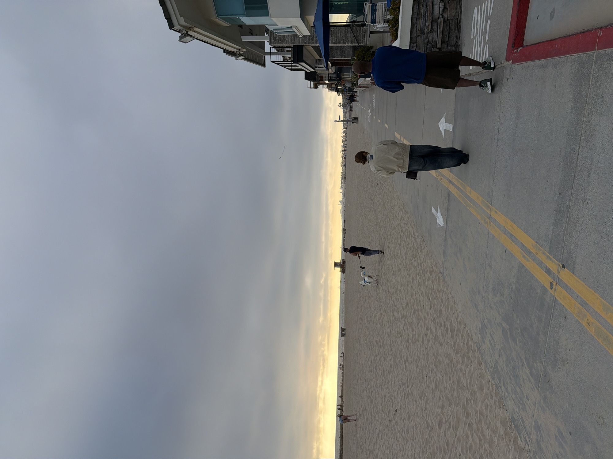 Here's a detailed description of the image:
**Location:** This image appears to be taken in Newport Beach, California, United States of America. The presence of the beach, buildings along the boardwalk, and the architectural style suggest this location.
**Scene:** The photograph depicts a beach scene during the day with cloudy skies. There's a stretch of sandy beach alongside a concrete boardwalk. Buildings are visible on the left side of the boardwalk.
**People:**
* Two people are walking on the boardwalk. One is wearing a blue shirt and dark shorts, and the other has a tan jacket and jeans.
* There are several other people on the beach, some with dogs.
**Objects:**
* The boardwalk has yellow lines dividing it.
* There are white arrow markings on the boardwalk.
* A lifeguard tower is seen in the distance on the beach.
**Actions/Interactions:**
* People are walking along the boardwalk and the beach.
* Some people are walking dogs.
**Environment:**
* The sky is overcast.
* The beach is sandy.
* Buildings are visible along the boardwalk.