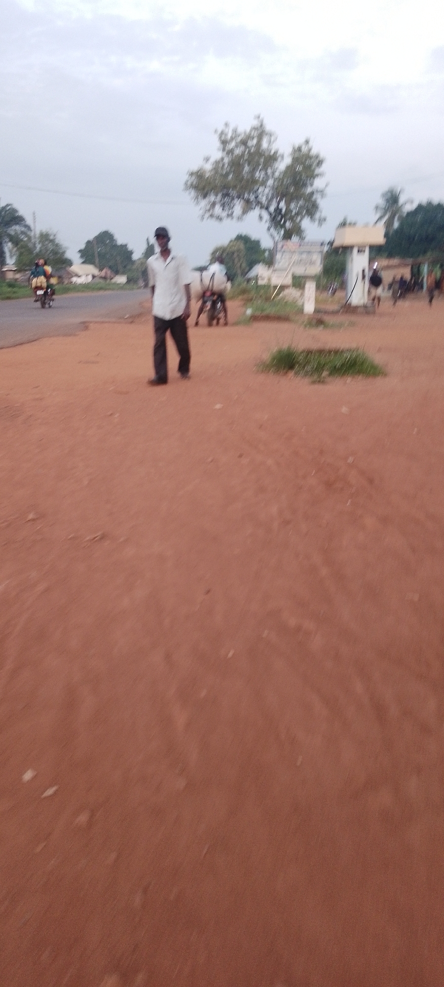 The image depicts a street scene in what appears to be the city of Ichol, Nigeria. The ground is covered in reddish-brown soil, suggesting a dry or dusty environment. In the mid-ground, a man is walking towards the viewer, wearing a light-colored short-sleeved shirt and dark trousers. He is also wearing a dark baseball cap. To the left of the man, a person is riding a motorcycle with a passenger behind them, also traveling towards the viewer. In the background, there are several buildings, trees, and what seems to be a utility pole with a sign. Another motorcycle is visible in the right background, with a person on it. The sky is overcast with a light grey color, indicating diffused light conditions. The overall scene suggests a typical day in a Nigerian city, with daily activities like commuting by motorcycle and people walking on the street.