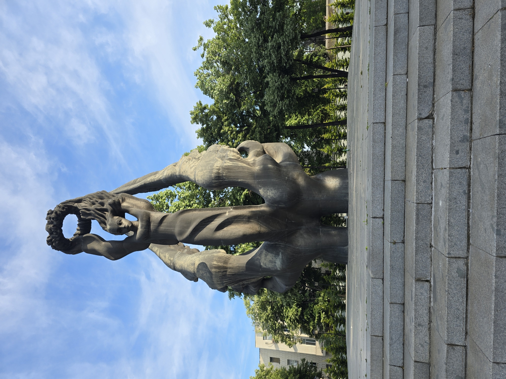 A statue stands in a city of Unknown, Unknown. The statue is constructed of dark grey stone, likely granite, and depicts a woman raising a laurel wreath above her head. She stands atop a base constructed of similarly-colored stone bricks. Behind the statue are green trees. The sky is blue with white clouds.