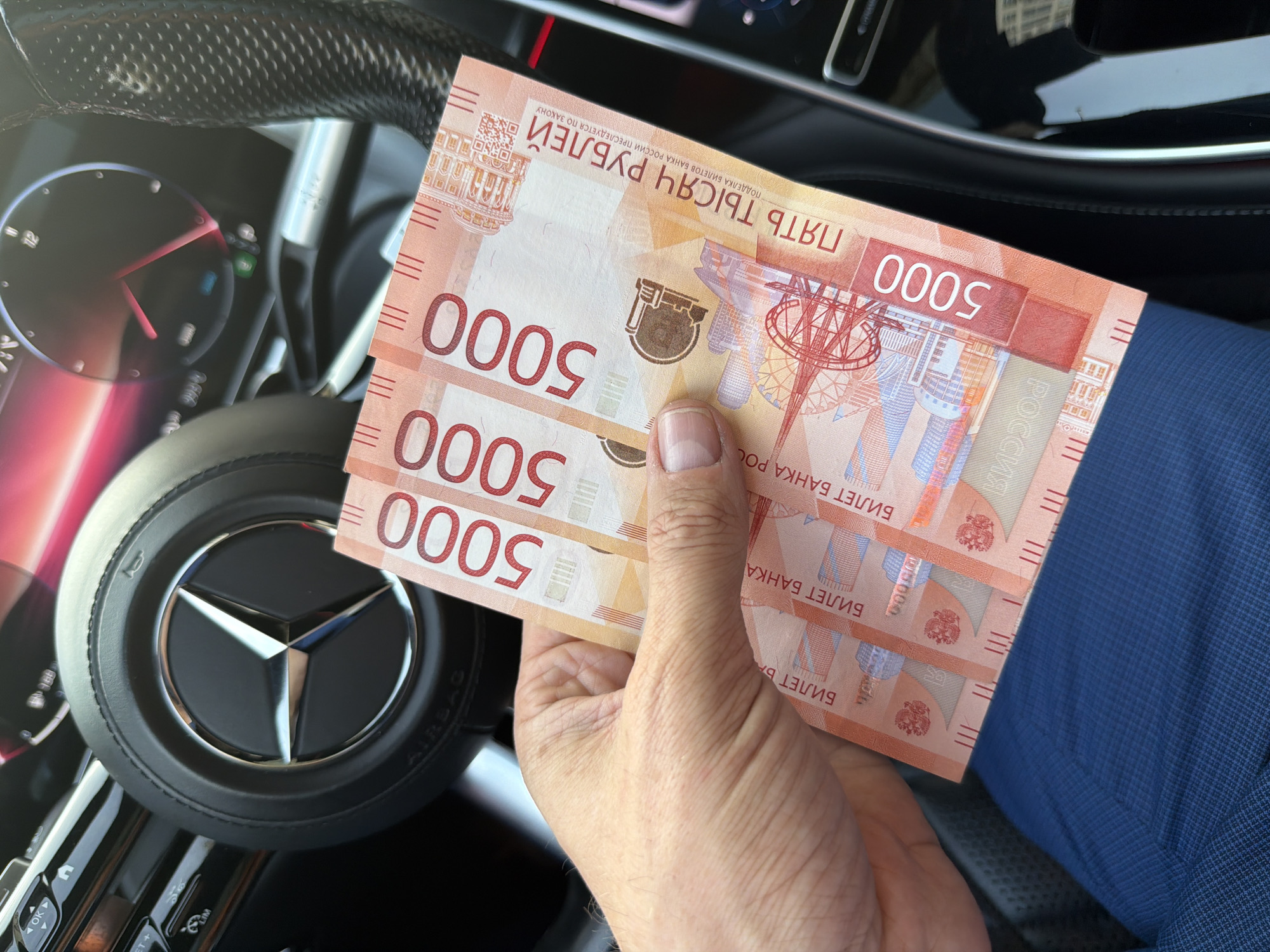 A hand holds three 5000-ruble banknotes, likely inside a car, near a Mercedes-Benz steering wheel. The banknotes are Russian currency. The blue fabric is the person's pants. The location is potentially in Khimki, Russia, based on the currency. The image suggests wealth.