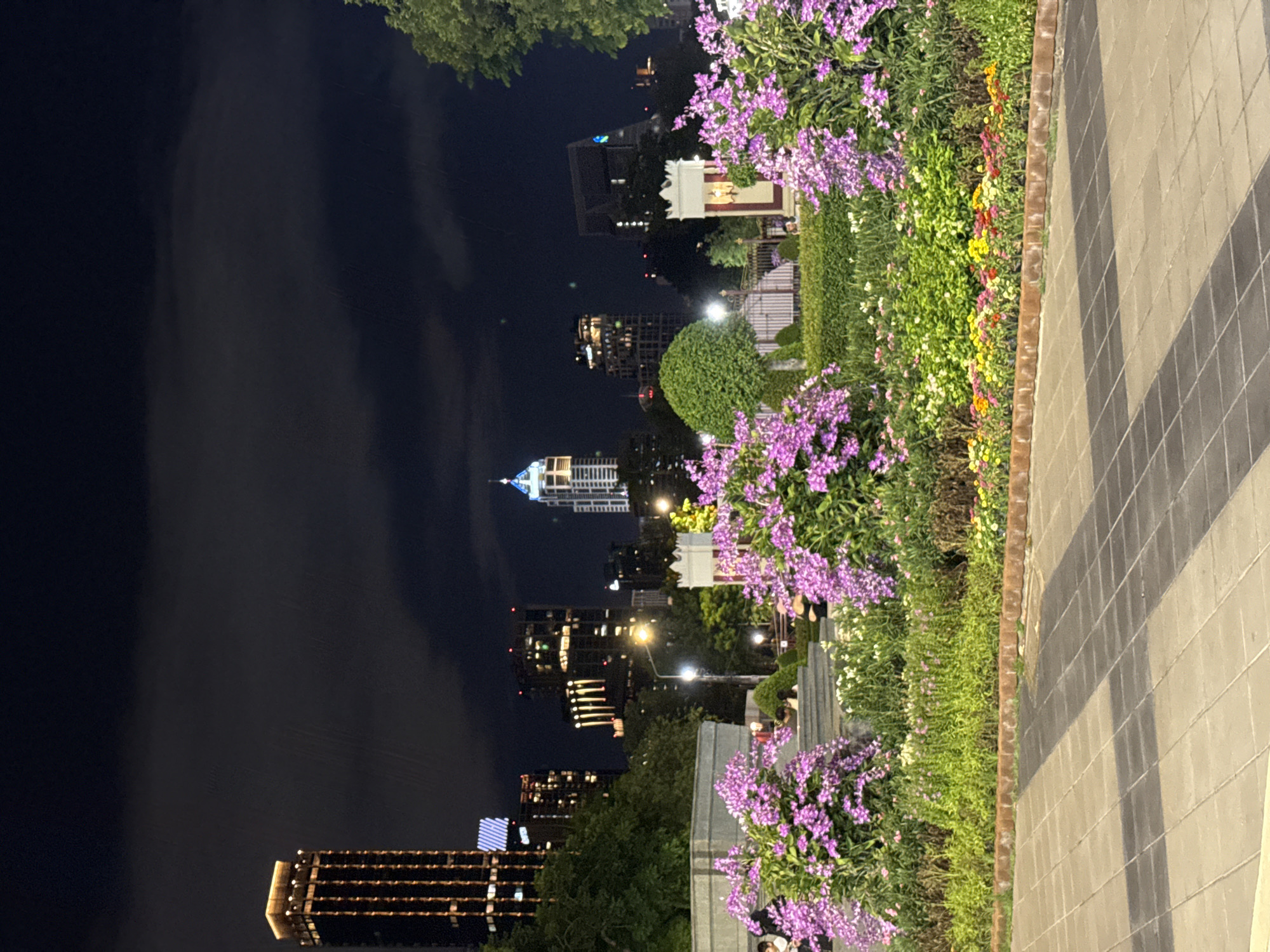 The image captures an evening scene in Bangkok, Thailand. In the background, multiple illuminated skyscrapers and high-rise buildings punctuate the night sky, situated across a body of dark water. Along the right side of the frame, a pedestrian walkway constructed with alternating gray and black tiles runs parallel to a meticulously maintained flower bed. Lush greenery, interspersed with vibrant purple and yellow flowers, creates a striking visual contrast to the urban backdrop. No individuals are visible in the foreground, suggesting a moment of quiet serenity amidst the bustling city.