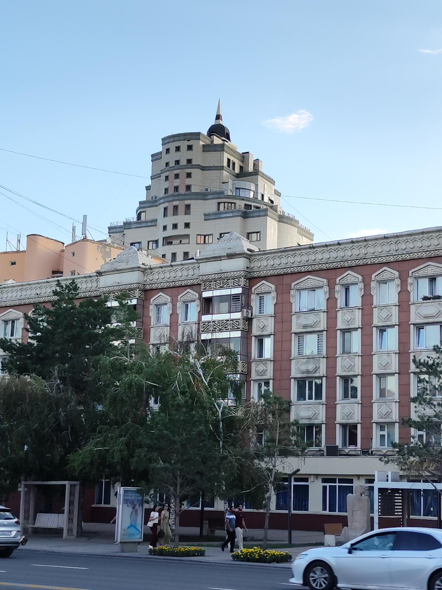 The image shows a multi-story building with a distinctive tiered upper section topped with a spire, located in Astana, Kazakhstan. The lower portion of the building is a reddish-brown color with ornate white trim around the windows. Trees are in the foreground, partially obscuring the lower floors of the building. On the sidewalk in front of the building, people are walking and standing near a bus stop shelter and a billboard. A white car is driving on the street in the foreground. The sky is clear and blue with a few wispy clouds.