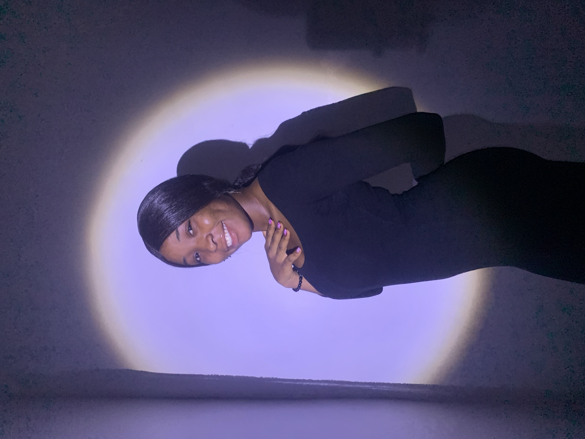 A medium shot photograph shows a dark-skinned woman wearing a black, long-sleeved dress. She is turned slightly to her left, looking at the camera and smiling. Her hair is styled straight back. She wears a bracelet on her left wrist, and her fingernails are painted pink. She has her left hand raised near her neck, with her fingers touching her neck. The background is a gradient of light and dark blue/purple. A circular, diffused light illuminates the area around the woman. The location in the city of Jos, Nigeria is not determinable from the image.