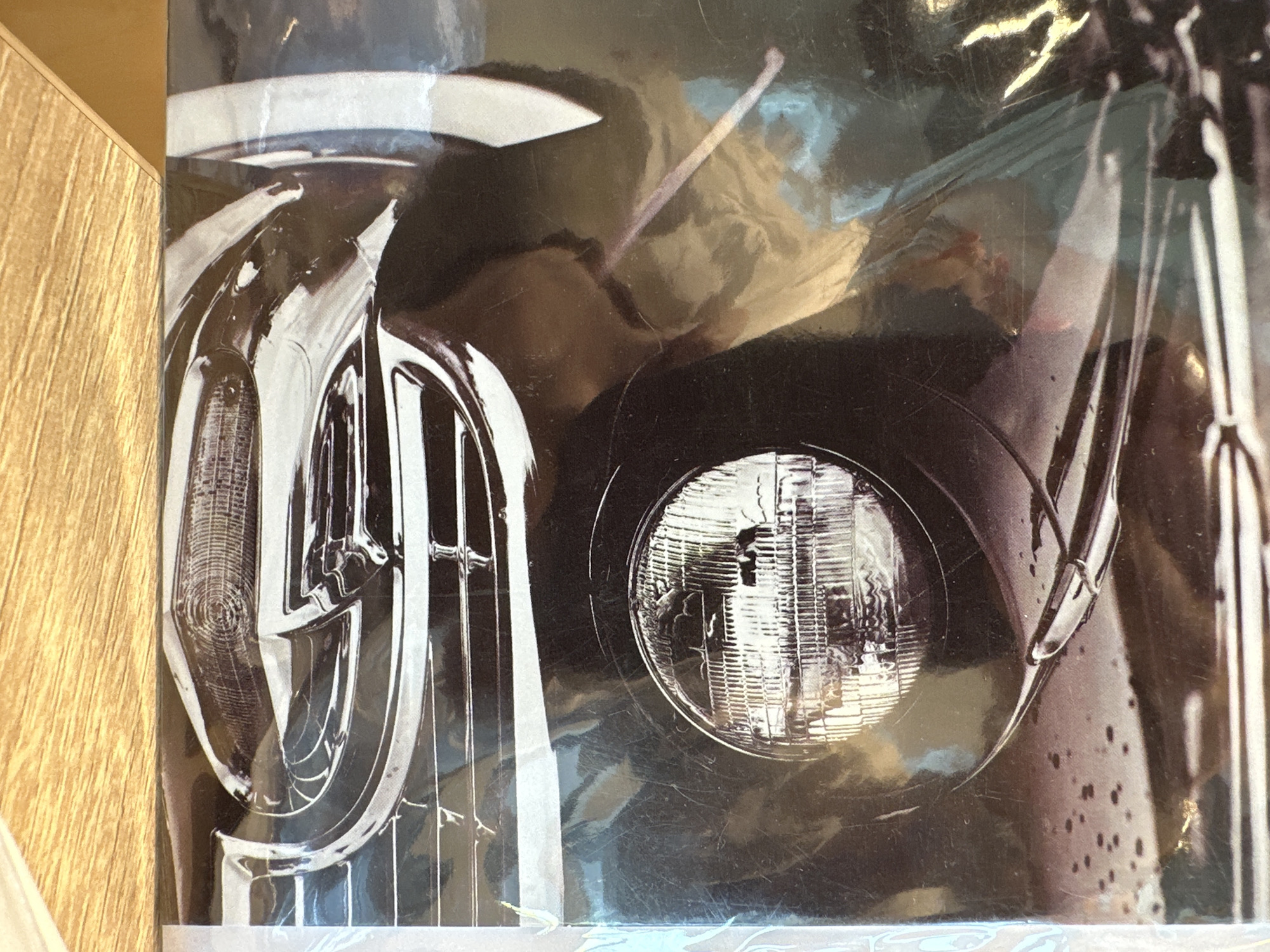 The image displays a close-up, angled view of the front right quarter of a dark-colored classic American automobile. The scene is located in Новоглаголево, Russia. A prominent circular headlight with a detailed fluted lens is centered in the upper-mid section. Below the headlight, a complex chrome grille structure with horizontal bars and vertical elements is visible, leading down to a wraparound chrome bumper. An amber-colored turn signal or parking light with a textured lens is integrated into the lower part of the bumper. The car's body features a dark, possibly black or deep blue, glossy finish, upon which numerous small water droplets are evident, suggesting rain or recent washing. Reflections of an unseen environment, featuring muted blue and brown tones, are visible on the car's painted surfaces and polished chrome. A chrome hood ornament or trim piece is present on the fender above the headlight. The vehicle's white sidewall tire is partially visible on the far left. The photograph itself appears to be a glossy print or poster, exhibiting surface scratches, dust, and significant reflections from an external light source, indicating it is covered by a protective layer or under bright lighting conditions. The bottom of the print rests on a light-colored wooden surface.