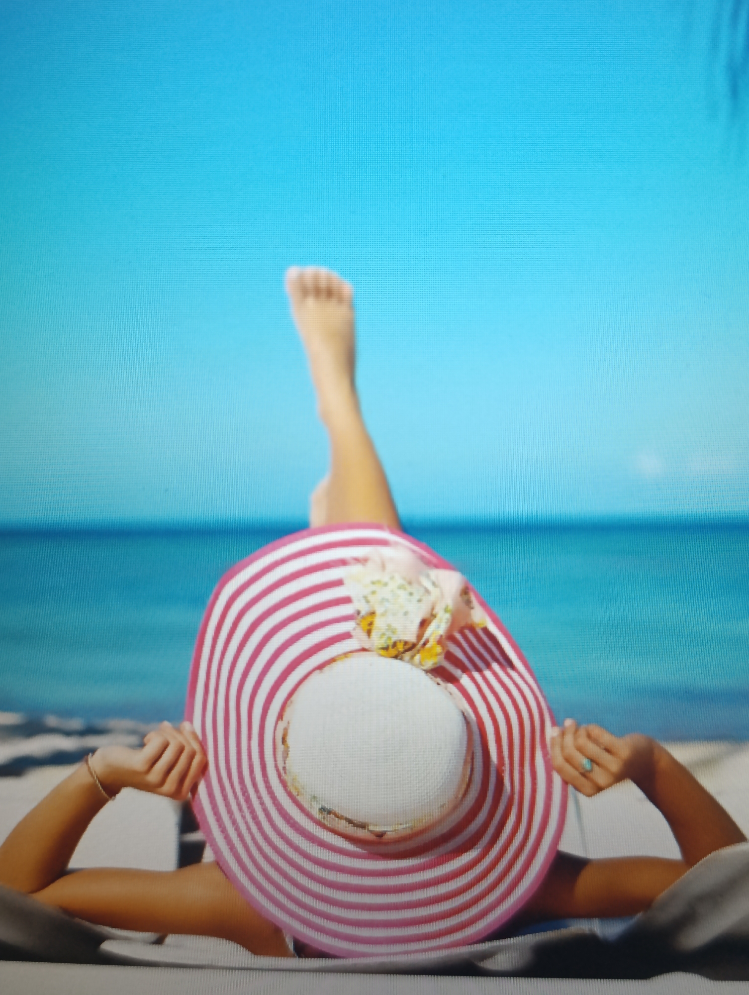 This media file shows a person from behind, lying on a beach chair with their legs extended upwards, against a backdrop of a bright blue sky and the ocean. The person is wearing a wide-brimmed hat decorated with pink and white stripes, and a colorful floral embellishment on top. They are also wearing a turquoise ring on one hand. The overall impression is one of relaxation and enjoyment of a sunny day at the beach. The time of day appears to be daytime, with clear skies and calm waters, suggesting pleasant beach weather. There is no visible text in the image.