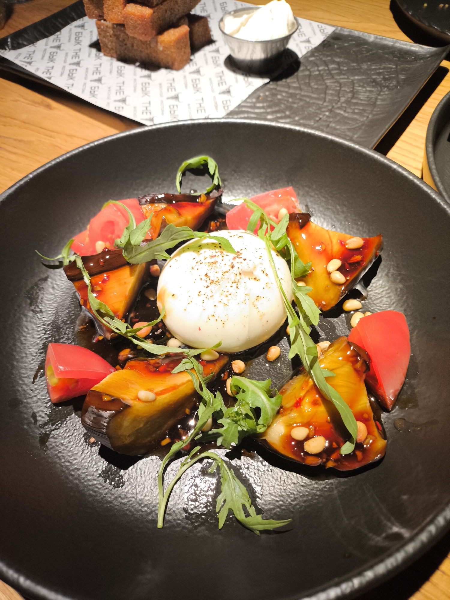 This is a close-up, overhead shot of a dish served in a black, textured bowl. The main subject is a salad composed of roasted eggplant wedges, bright red tomato pieces, and a central ball of what appears to be burrata cheese. The dish is drizzled with a dark, glossy sauce and sprinkled with pine nuts and fresh green arugula leaves. The lighting suggests an indoor setting, possibly a restaurant, with a warm wooden surface visible beneath the bowl and in the background.
In the upper portion of the image, partially visible, are stacked pieces of dark bread and a small metal container holding a white, creamy substance, likely a dip or spread. These items are placed on a black placemat with a repeating logo or text pattern. The visible text, in a stylized font, appears to be "THE BYK" in English and possibly a Cyrillic transliteration as well ("БЫК" which translates to "Bull"). The overall presentation is appetizing, highlighting the textures and colors of the ingredients. There are no people or animals visible in the frame. The time of day and weather are not discernible.