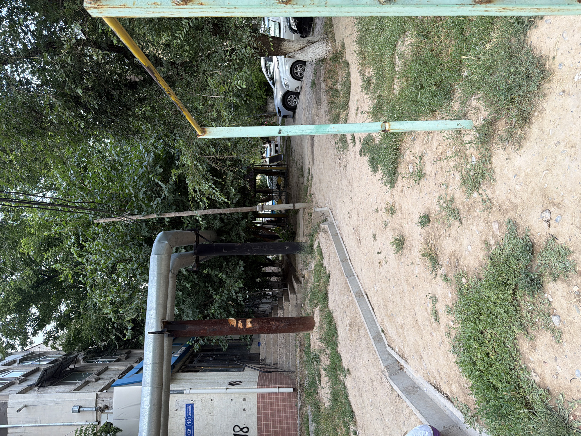 This media file captures an outdoor scene in what appears to be a residential area, possibly a courtyard or alleyway, with a hint of urban decay. The image is oriented sideways, with the ground running vertically on the right side of the frame and the sky at the bottom.
The main subjects are several large, mature trees with dense green foliage, which dominate the left side of the image and cast shadows. Alongside the trees, there is a section of a building, showing some architectural details like windows and pipes. A prominent feature is a set of large, insulated pipes running horizontally across the middle ground, supported by rusty metal structures.
In the background, a dirt path or unpaved road winds its way between the trees and buildings. There are cars parked along this path, suggesting it is a functional area for residents. One car, a white SUV, is clearly visible, positioned between tree trunks. Further down the path, more cars and what looks like a covered seating area or small kiosk can be discerned.
The ground is a mix of sandy soil, patches of dry grass, and scattered stones. A concrete curb or drainage channel runs along the edge of the path on the right side. A metal structure, possibly part of a fence or playground equipment, with a faded green and yellow paint scheme, crosses the upper portion of the image.
Notable details include the rust on the metal supports for the pipes and the weathered appearance of the metal structure. The lighting suggests it might be daytime, perhaps late afternoon, given the shadows. There is no clear indication of specific weather conditions, but the greenery implies it's not a harsh season.
Visible text includes a blue and white sign on the building with Cyrillic lettering and the number "18" in a circle. Another instance of the number "18" is printed in a larger font on the building's facade.
The overall impression is one of a lived-in, slightly neglected urban environment, with elements of nature interspersed with man-made structures.