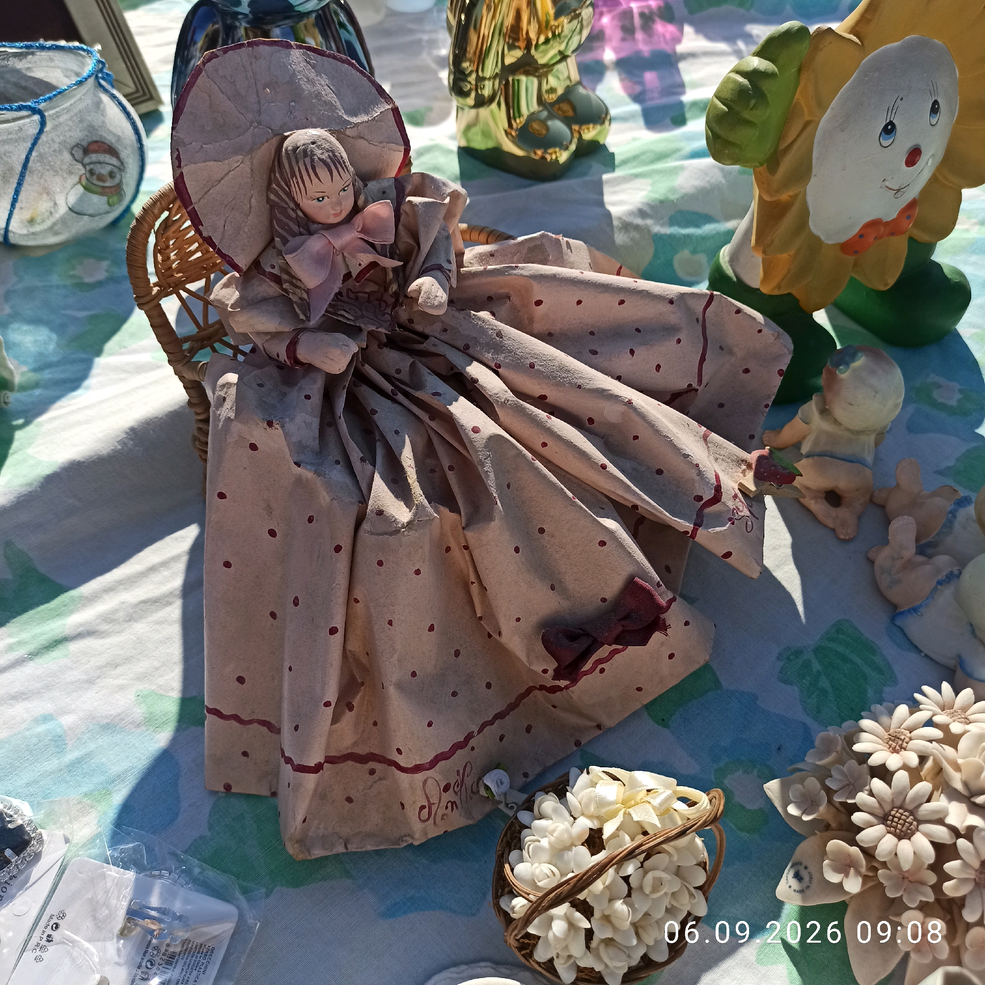 This image shows a close-up, eye-level view of a doll in a woven chair, displayed on a tablecloth. The doll is dressed in a large, pale pink polka-dot dress and a matching hat. The tablecloth has a blue and green floral pattern. In the background, there are other decorative items, including a glass vase with a snowman design, a golden statue, and a ceramic sunflower. To the right of the doll, there are small ceramic baby figures. In the foreground, there is a small woven basket filled with white flowers and another cluster of white ceramic flowers. The scene is set outdoors, as indicated by the sunlight and shadows. The date and time "06.09.2026 09:08" are superimposed on the image. The location is stated as Sannicandro di Bari, Italy, though no specific landmarks are visible to confirm this.