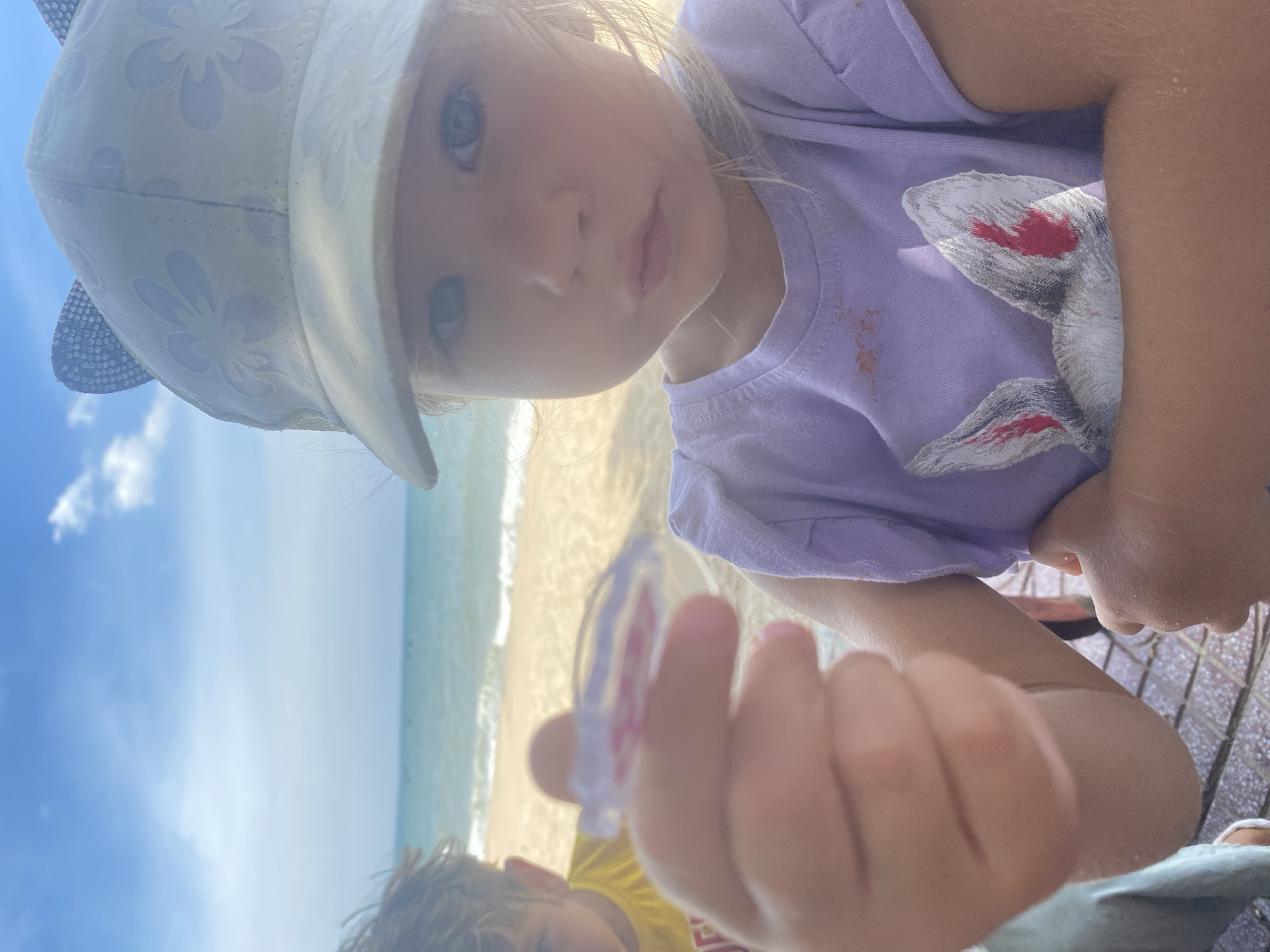 A close-up, waist-up shot captures a young Caucasian girl wearing a white baseball cap with a floral pattern and cat ears, a purple t-shirt with a rabbit graphic, and standing in front of a beach scene. She is holding a clear plastic object with pink inside up to the camera with her left hand. Her expression is neutral. To the left of the frame, a young Caucasian boy wearing a yellow t-shirt is partially visible. The background shows a sandy beach, turquoise ocean, and a bright blue sky with scattered clouds. The location is Phu Thuy Ward, Vietnam.