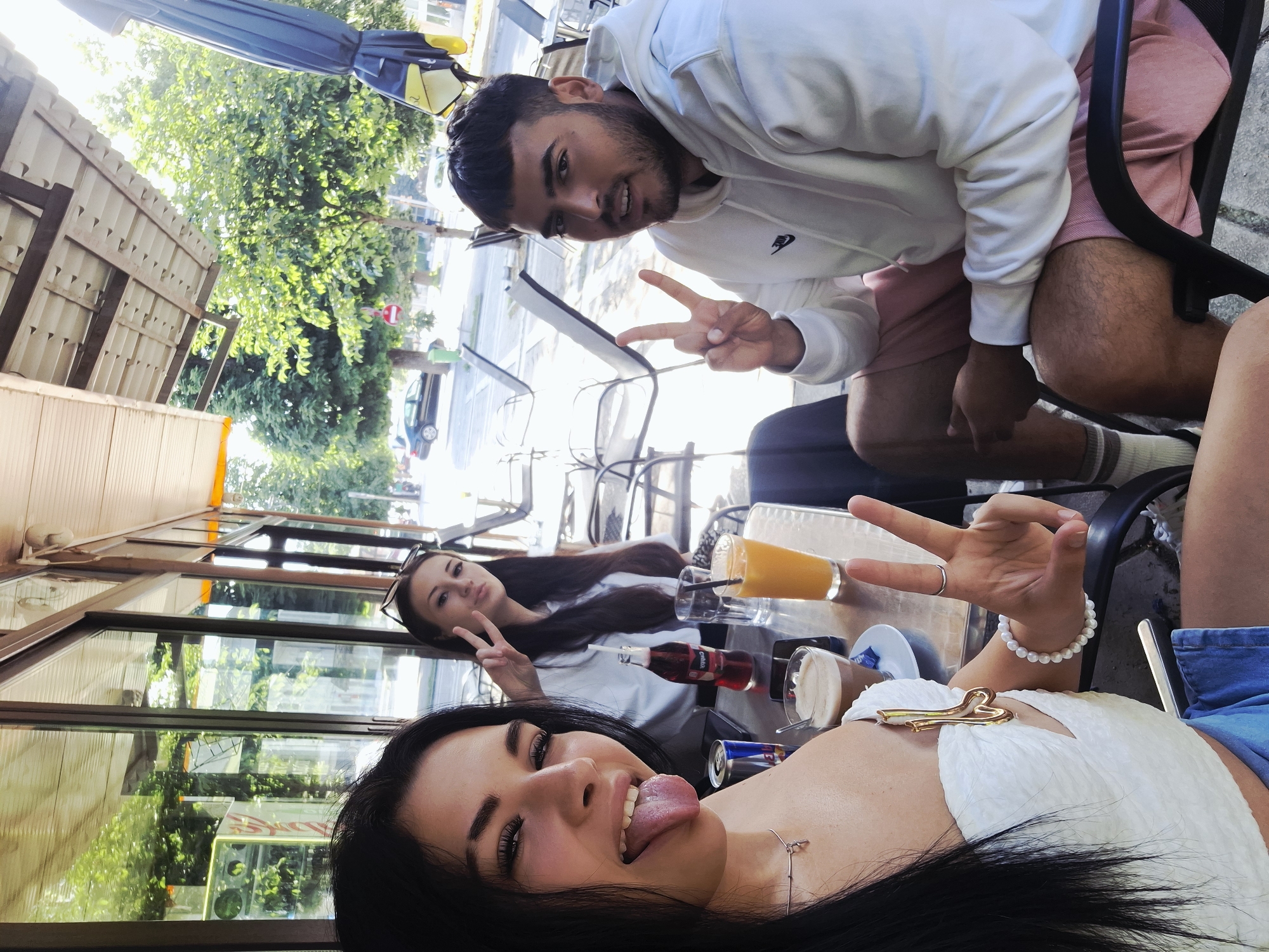 In Mezdra, Bulgaria, three individuals are seated at an outdoor cafe. A woman in the foreground, with long black hair, is sticking her tongue out at the camera, wearing a white top. To her left, another woman with long brown hair is making a peace sign. A man with dark hair and a beard, wearing a white hoodie and pink shorts, is seated to the right, also making a peace sign. On the table, there are drinks including an orange juice, a coffee, and a bottle of red soda, along with a can of soda. The cafe is situated outdoors, with trees and a street visible in the background.