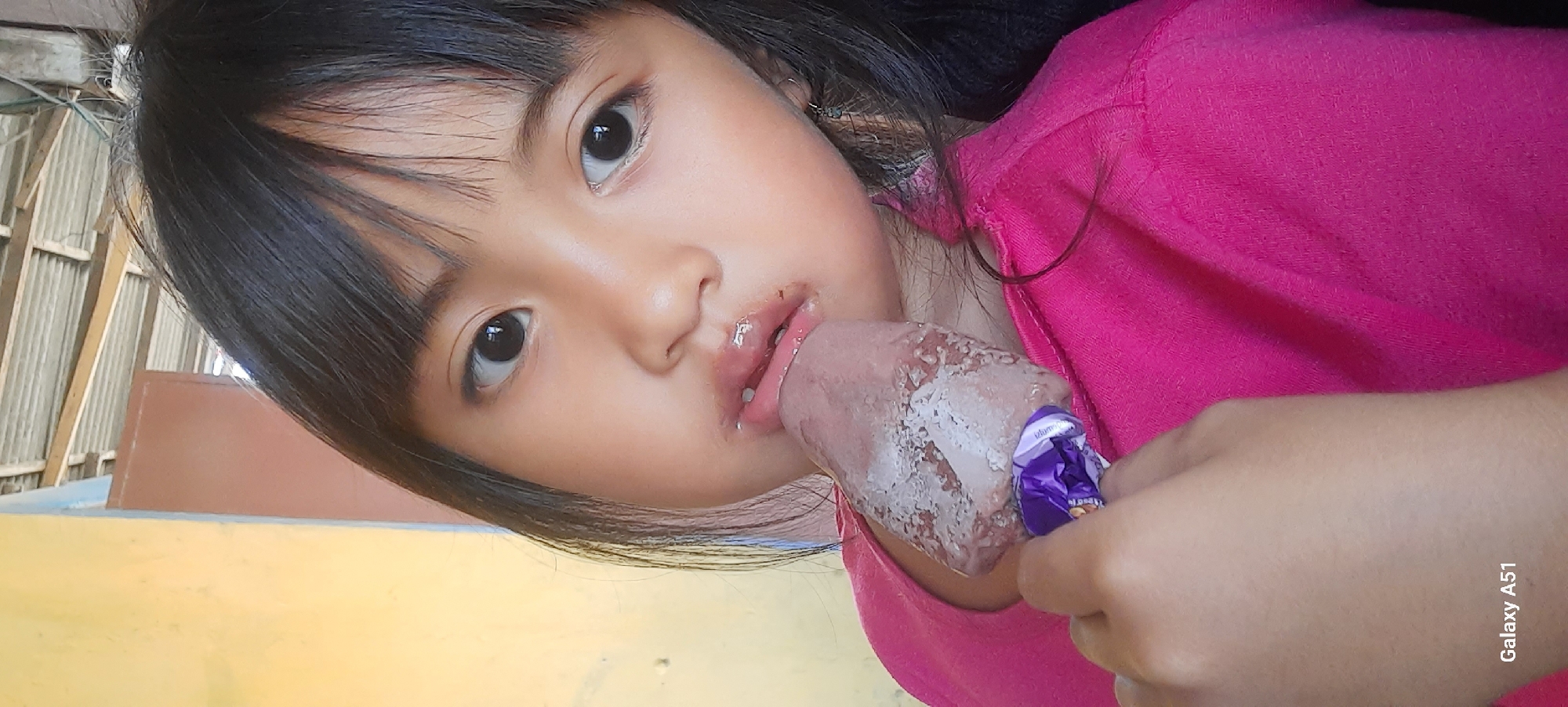 The media file shows a close-up, horizontally oriented shot of a young girl in Selopuro, Indonesia, enjoying an ice cream bar. The girl, with dark hair and dark eyes, is wearing a bright pink shirt. She is holding the ice cream bar with her right hand, and her tongue is extended, licking the partially melted treat which is covered in white remnants. Chocolate residue is visible around her mouth.
The background appears to be outdoors, with corrugated metal fencing and wooden structures suggesting a rustic or unfinished building. A yellow surface is visible in the foreground. The lighting suggests it might be daytime, but no specific time of day is discernible. The girl's expression is focused on the ice cream. The visible text "Galaxy A51" indicates the device used to capture the image.
