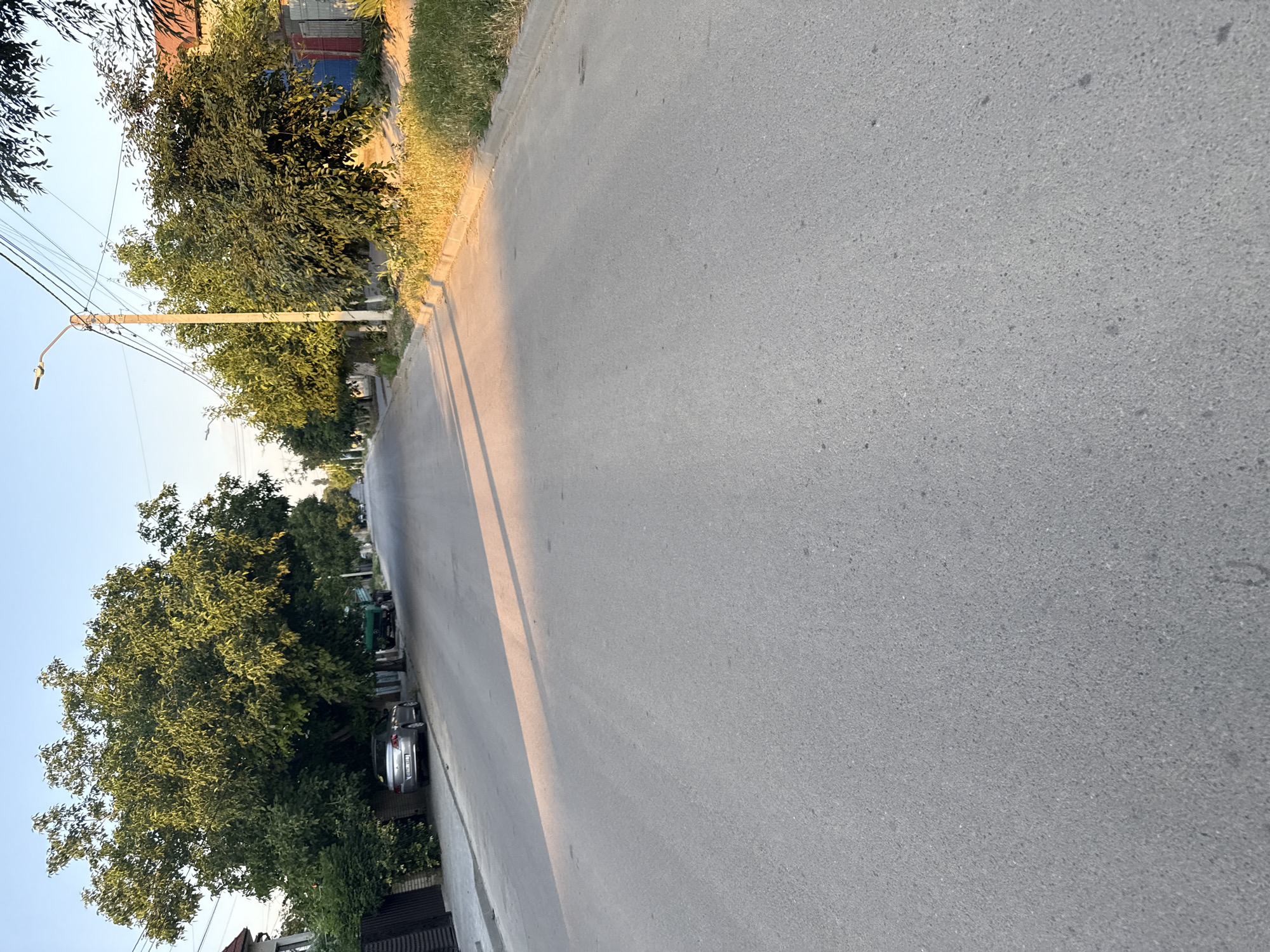 The image depicts a street scene in Cahul, Moldova, under a clear sky at what appears to be either sunrise or sunset, indicated by the long shadows cast across the asphalt road. The road itself is paved and shows signs of wear. On the left side of the road, a silver car is parked. Further down the street, a green truck is visible. The sides of the road are lined with trees and vegetation, with fences and buildings belonging to residential properties. A street lamp stands on the right side of the road. The scene is tranquil, with no people or active interactions visible.