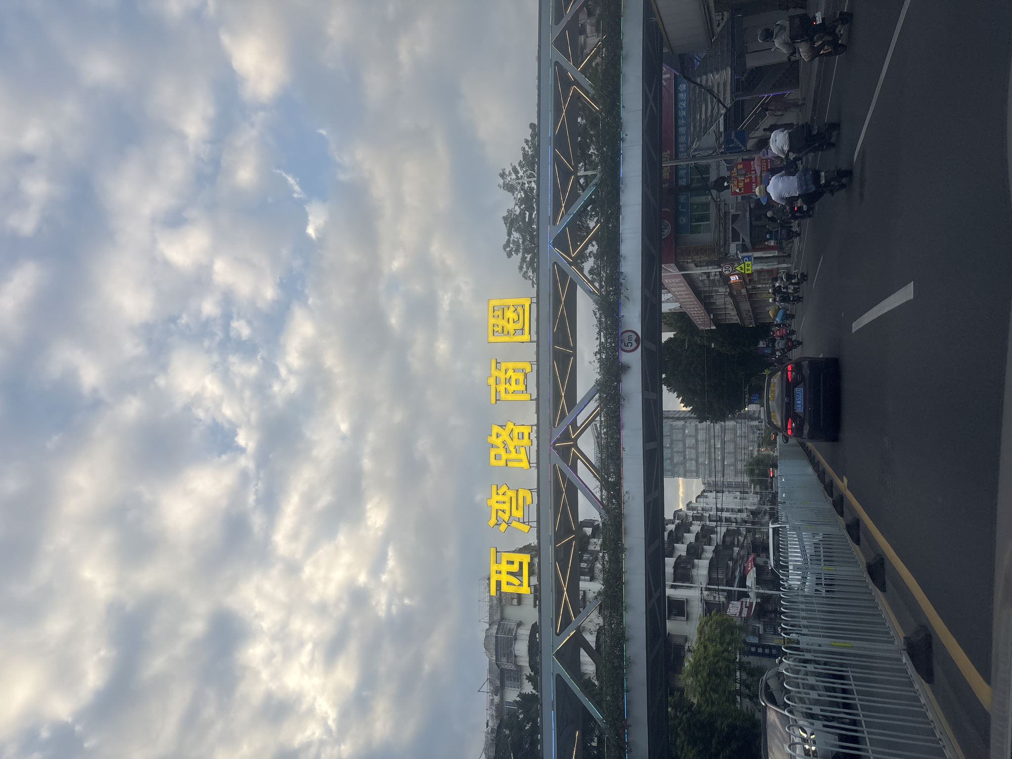 The image depicts a street scene in Liwan District, China, under a cloudy sky. A pedestrian overpass spans the road, adorned with decorative lights and greenery. Prominently displayed on the overpass are large, illuminated yellow characters that read "西湾路商圈" (Xī Wān Lù Shāng Quān), translating to "Xiwanshan Road Business Circle." Below the overpass, traffic flows along a multi-lane road. Several vehicles, including cars and numerous motorcycles, are visible. Some motorcycles are carrying passengers, and one driver is wearing a white shirt and a blue bag on the back of their vehicle. On the sidewalks, buildings with various architectural styles line the street, some appearing older and others more modern, including a tall, slender skyscraper in the background. A speed limit sign indicating "5m" is visible on the overpass support. Further down the road, commercial signs are displayed on the buildings, and pedestrians are walking along the sidewalk. The overall atmosphere suggests a bustling urban environment during daylight hours.