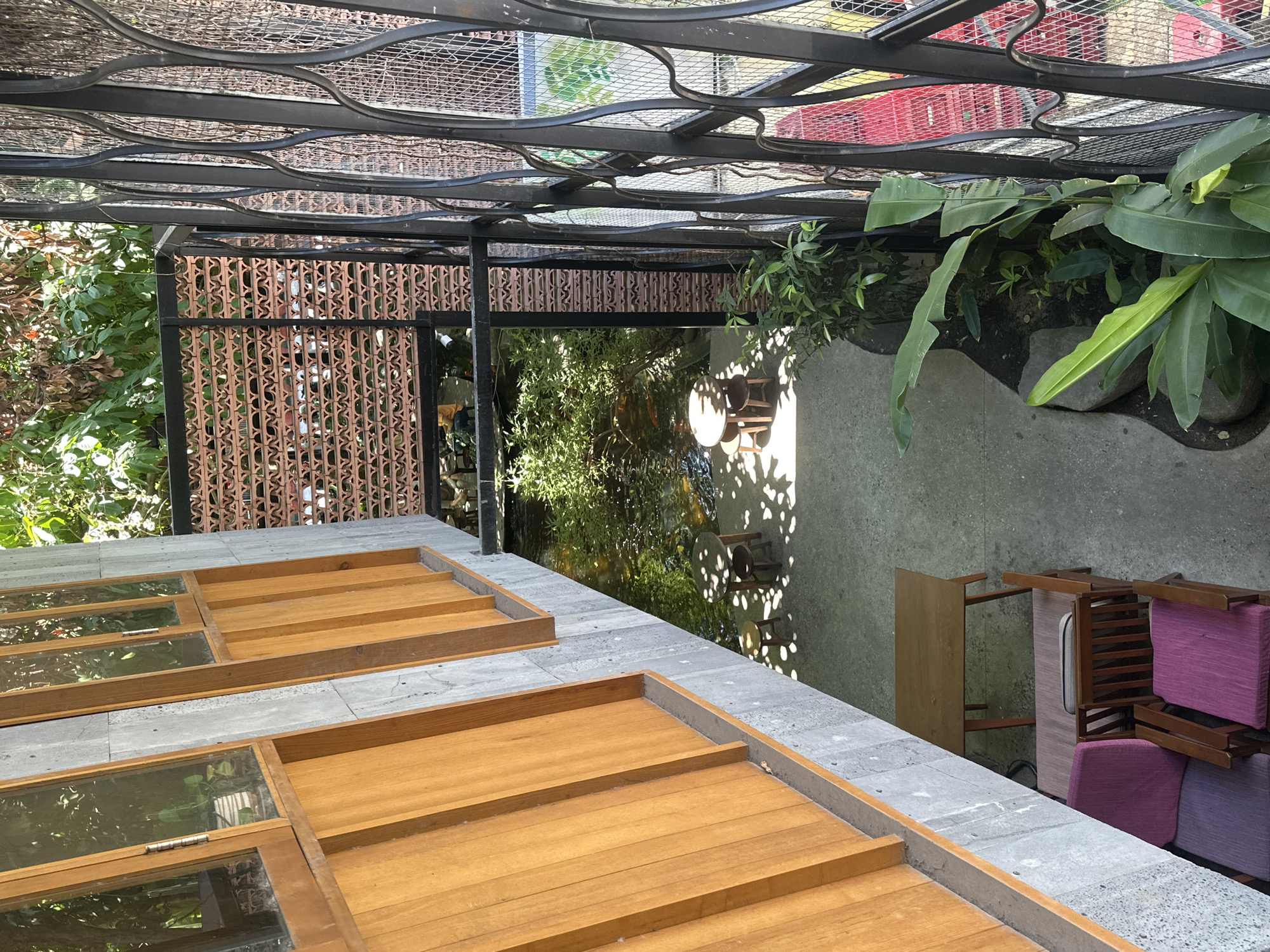The image captures an outdoor courtyard area in Đà Nẵng, Vietnam, with a distinctive architectural style blending concrete, wood, and metalwork. In the foreground, to the left, a wooden structure with several glass windows is visible, featuring prominent wooden beams. Adjacent to this building is a small pond filled with lush greenery and visible koi fish. Two small wooden tables with accompanying stools are placed around the pond, casting shadows on the concrete ground. To the right of the pond, a decorative metal fence with an intricate pattern encloses the space, revealing a glimpse of what appears to be a red structure beyond. Further to the right, large-leafed plants grow in a planter. In the bottom left of the image, a set of cushioned chairs, one purple and one pink, are arranged around a low wooden table. The scene suggests a tranquil, outdoor seating or dining area, possibly within a cafe or restaurant, designed to integrate nature and modern aesthetics.