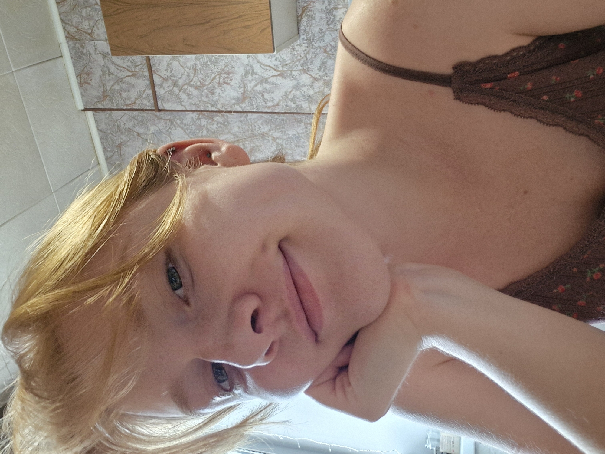 A selfie taken from a low angle shows a fair-skinned woman with blonde hair. She is wearing a brown floral-patterned top with a thin strap. Her left hand is resting under her chin. The background includes a cabinet with a wood-grain door and tiled walls. The location is Unknown, Unknown.