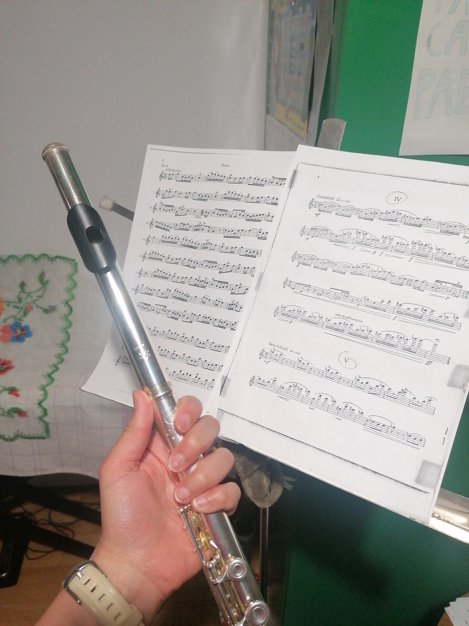 The image shows a person holding a flute in their left hand. A piece of sheet music is displayed on a music stand in the background. The sheet music is white with black notes and text. The background behind the music stand is a combination of colors, including green and white. The environment appears to be indoors.