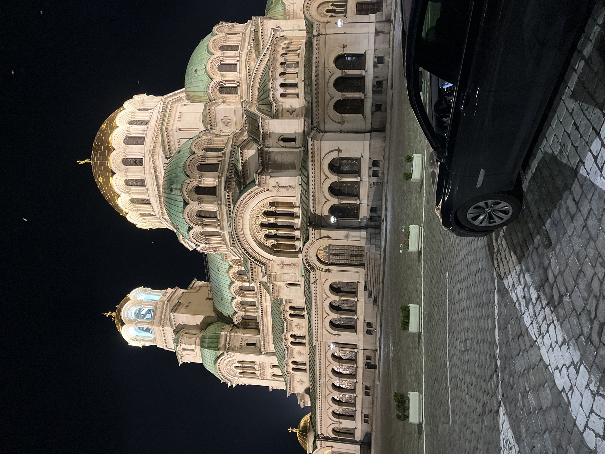 The image shows the Alexander Nevsky Cathedral in Sofia, Bulgaria, at night. The cathedral is brightly lit, highlighting its architectural details, including the domes with green and gold accents. A dark-colored car is parked on the right side of the frame. The ground is paved with cobblestones, with planters positioned along the edge of the pavement. The sky is dark, indicating it is nighttime. There are no visible people in the frame. The overall scene is static and focuses on the architectural grandeur of the cathedral and the car parked beside it.