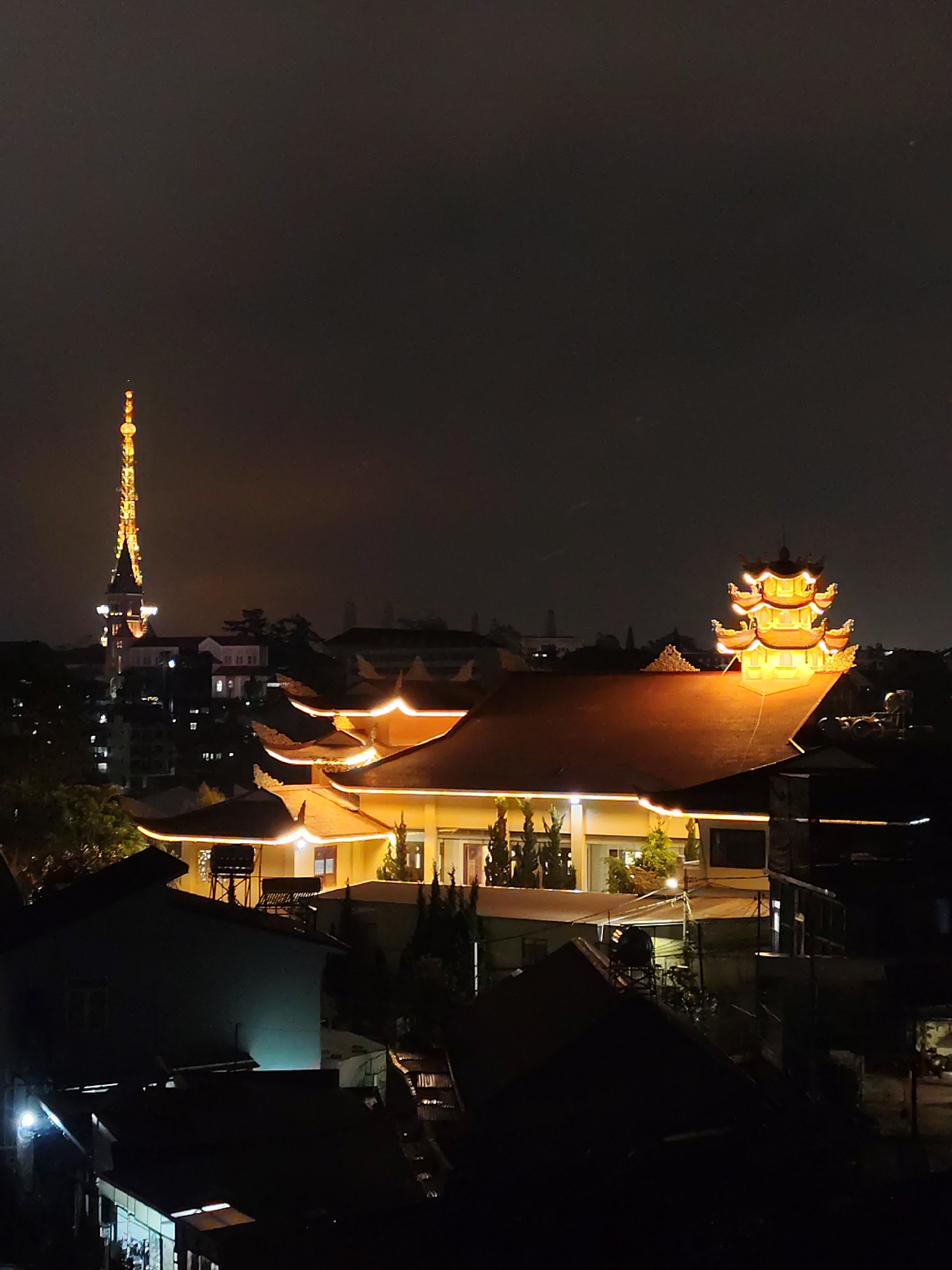 An elevated nighttime view in Da Lat, Vietnam reveals a cityscape dominated by illuminated structures. In the foreground, a multi-tiered building with a red tile roof is prominently lit, its edges defined by rows of lights. Adjacent to it are darker, lower buildings with sloping roofs, partially obscuring details of the surroundings. In the background, a tall, slender, brightly lit tower stands out against the night sky. Other buildings are visible, forming a skyline with scattered lights. The overall environment suggests a densely populated urban area at night.