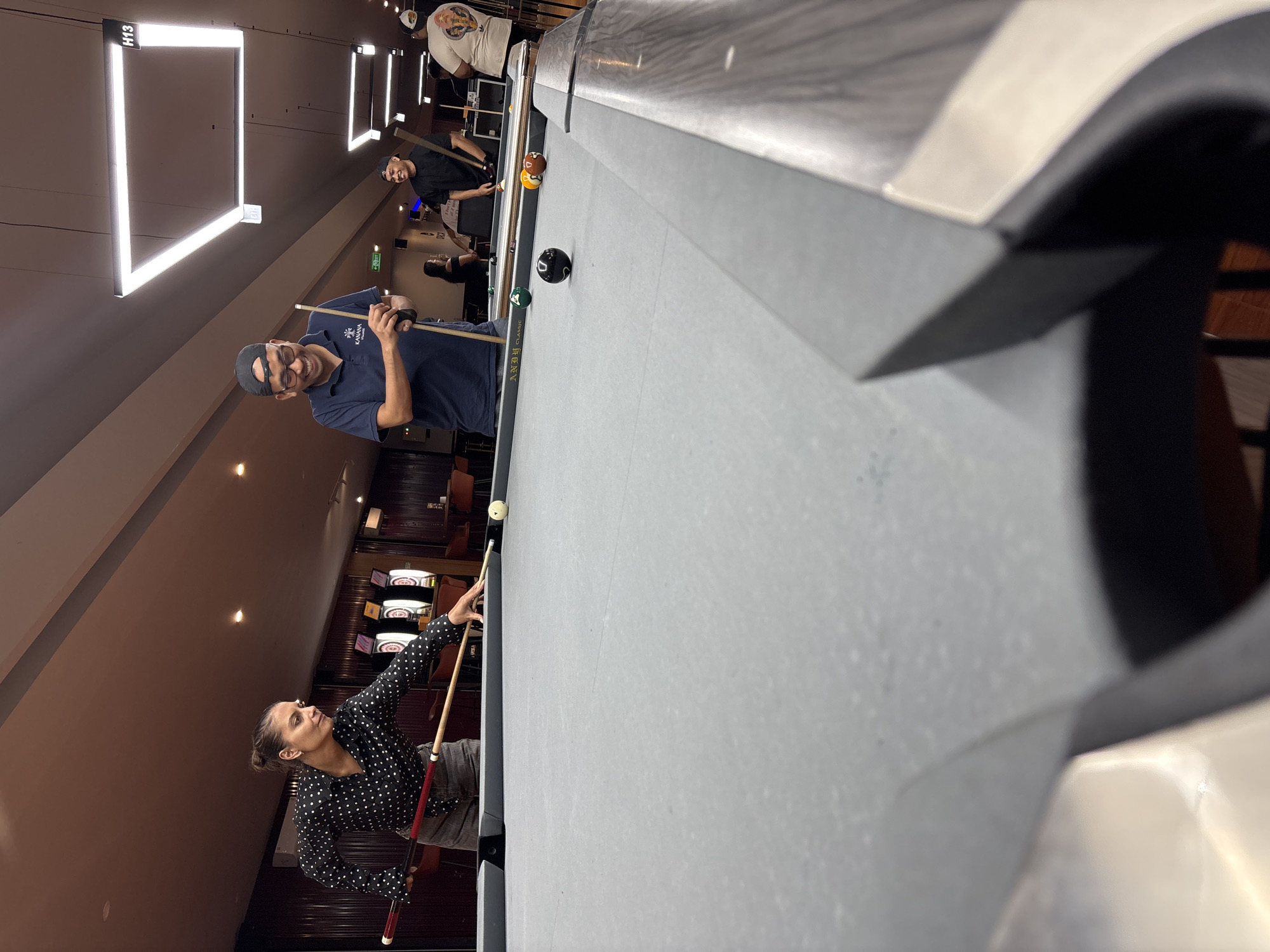 In Kuta Selatan, Indonesia, a group of people are gathered around a billiard table in an indoor establishment. In the foreground, a woman wearing a black and white polka dot shirt and grey shorts is positioned to strike a billiard ball with a cue. The billiard table is covered in a light grey cloth and several balls are visible, including a black ball, a green ball, and an orange and brown striped ball. Behind her, a man in a blue polo shirt and a black baseball cap is holding a cue, smiling and looking towards the table. Further in the background, other individuals are present, some also holding cues. The environment appears to be a leisure or entertainment venue, with dimly lit ceilings and modern light fixtures. Dartboards are visible on the wall in the background.