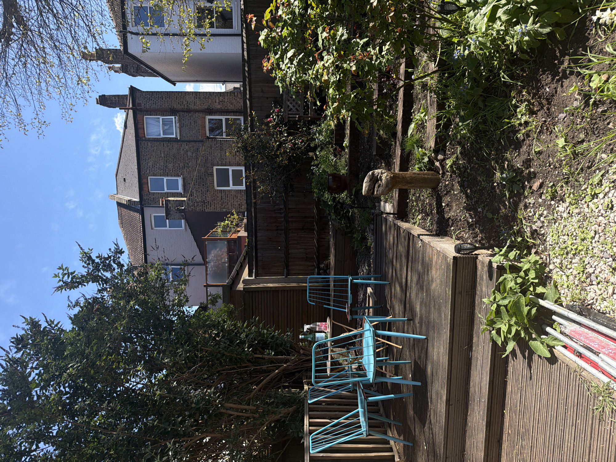 The image shows a garden in London, United Kingdom. The scene features a wooden deck with a set of blue chairs and a small table. On the table are several glass bottles. The deck leads to a garden with plants and a wooden fence. Behind the garden is a brick building with multiple windows and a light-colored building beside it. A tree occupies the lower left of the frame. Ladders lay on the right of the picture. The sky is blue with few clouds.