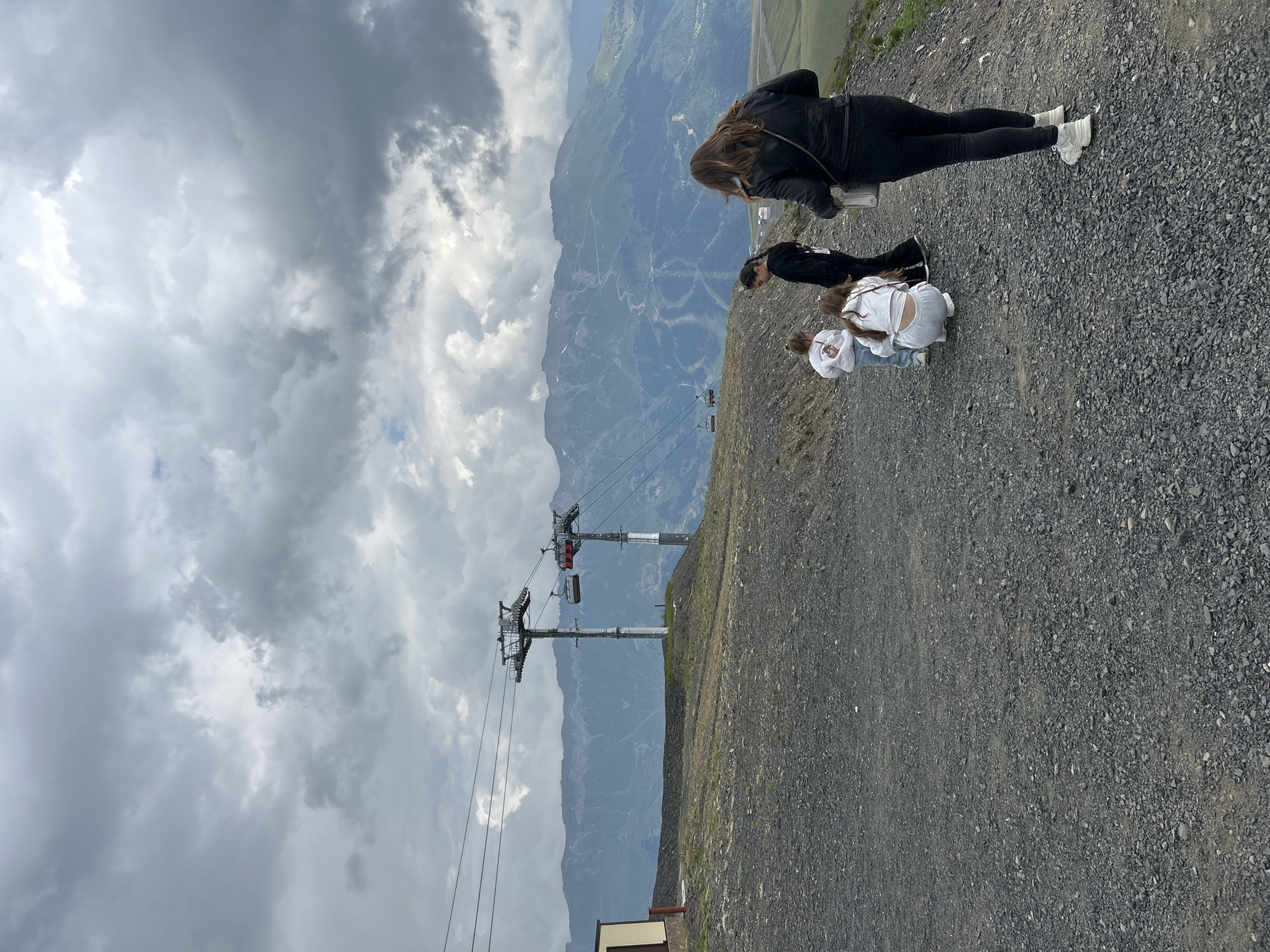 In Lunnaya Polyana, Russia, on a cloudy day, four individuals stand on a gravelly slope overlooking a mountain range. The foreground is dominated by the rough, dark gravel. In the mid-ground, two ski lift towers with cabins are visible, strung between cables. A building with a yellow facade is situated to the left. The background features a vast expanse of mountains, with visible ski tracks winding down their slopes. Three young girls and one adult woman are positioned in the scene, with two girls crouching and the other two standing. The woman in the foreground, viewed from behind, wears dark clothing and white sneakers. The sky is filled with heavy, gray clouds.