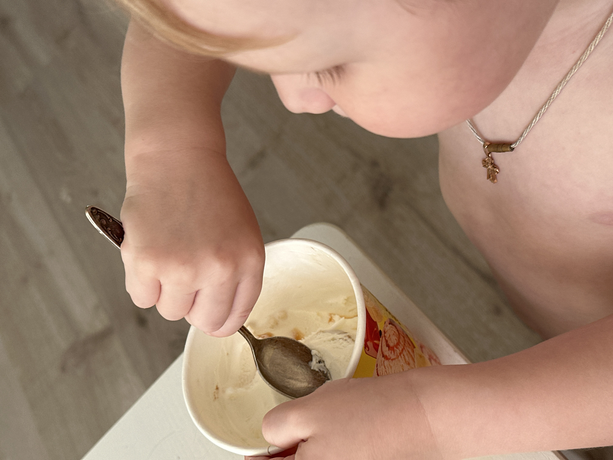 A toddler is shown from an overhead perspective. The toddler is fair-skinned with blonde hair, and is wearing a necklace with a cross pendant. The toddler is holding a metal spoon in their left hand, which is inside a partially empty paper container of ice cream. The container is being held by the toddler's right hand. The background is a light-colored wooden floor and a white table. The location of Votkinsk, Russia is not evident from the image.