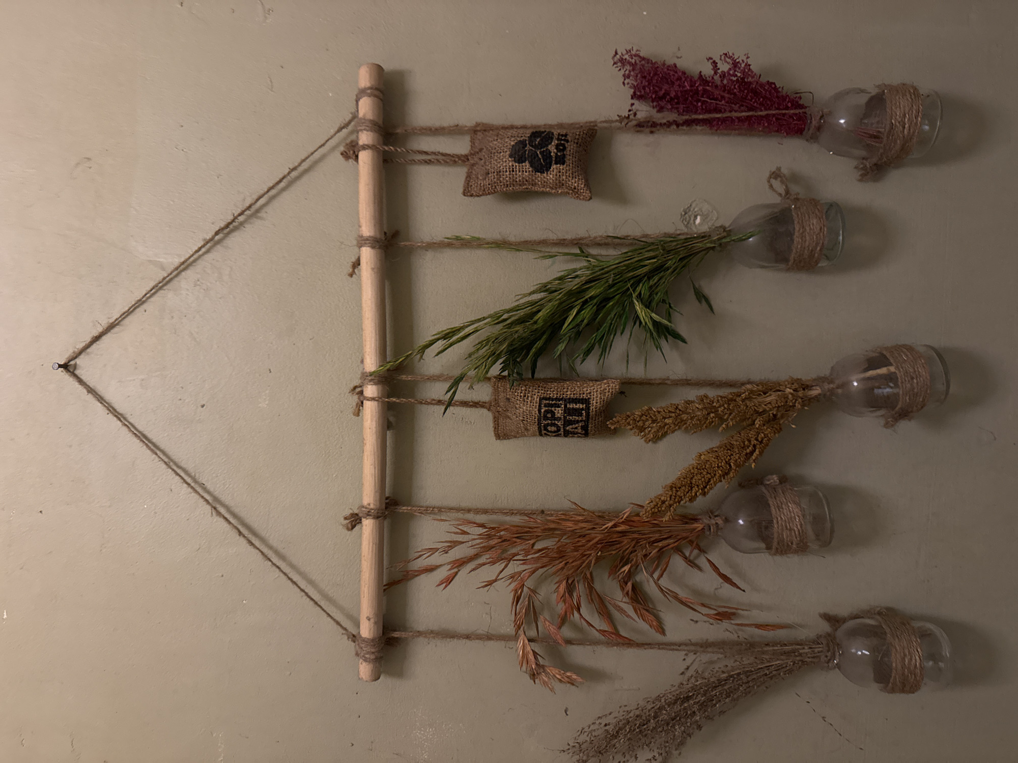 This is an eye-level, well-lit photograph of a rustic wall-mounted decoration set against a plain, light-colored wall, likely in an indoor setting. The main structure consists of a vertical, light-toned wooden dowel or rod, acting as a central support. Twine or rope is intricately wrapped around this dowel at several points, extending horizontally to create a ladder-like effect, and also forming a triangular support structure on the left, anchored by a nail or hook.
Attached to the horizontal twine strands are five clear glass bottles or vases, each wrapped with twine around its neck, giving them a handcrafted appearance. Each bottle holds a distinct bunch of dried flora or grasses. From top to bottom, these include: dark red/purple textured dried flowers, vibrant green leafy grasses, light brown seed-head-like dried sprigs, russet-orange feathery plumes of dried grass, and delicate light brown/greyish fine-textured dried grasses.
Two small, rectangular burlap sacks are also incorporated into the design. The top sack features a black paw print logo and partially visible text that appears to read "SOIL". The lower burlap sack displays the text "KOPI ALI" in black. The overall aesthetic is natural, earthy, and artisanal. The lighting suggests it's daytime indoors or under warm artificial light, with subtle shadows cast on the wall behind the objects. Given the "Kopi Ali" text and the location context of Kuta Selatan, Indonesia, this decoration might be found in a cafe, shop, or home, reflecting local elements and a natural theme.