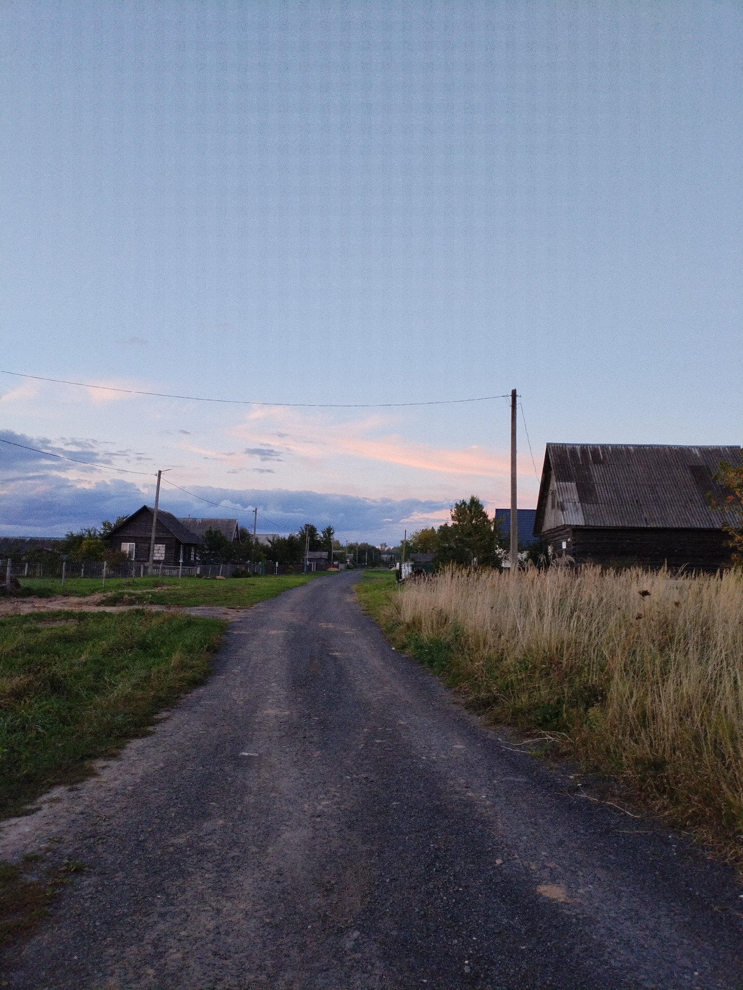 The image depicts a rural dirt road in the city of Bialynichy, Belarus. The road is unpaved and appears to be made of gravel and dirt. On either side of the road are grassy areas and dry, tall grass. Several wooden houses are visible in the background, consistent with a village or rural setting. Utility poles with wires are present. The sky is a pale blue with some wispy clouds tinged with pink, suggesting it is either early morning or late evening. There are no people or animals visible in the image. The overall scene is one of a quiet, somewhat undeveloped rural landscape.