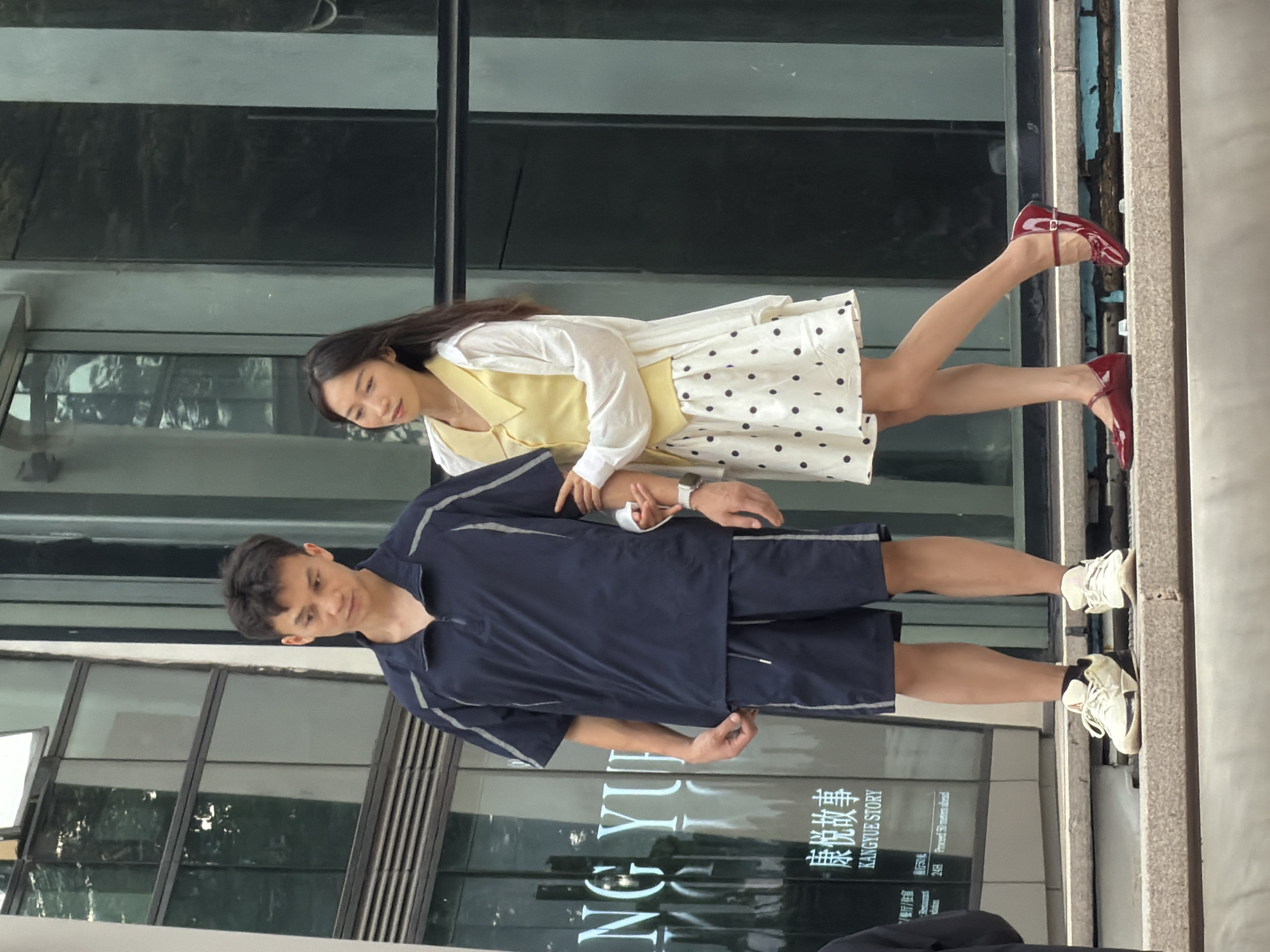 Two individuals, a man and a woman, stand on a stepped, grey concrete ledge in front of a building with large reflective glass windows. The man, on the left, wears a dark navy blue collared shirt with short sleeves and grey piping, and matching dark navy blue shorts. He has black hair and is looking down and to his left. The woman, on the right, has long dark hair and wears a white long-sleeved shirt over a light yellow sleeveless vest. Her white skirt has black polka dots. She is wearing red ballet flats with a strap and bow detail. The man's right arm is extended slightly, and the woman's left hand is touching his right arm. A sign on the building behind them displays Chinese characters and the text "KANGYUE STORY". The environment suggests an urban setting, specifically in the Yuzhong District of China, given the signage. The lighting indicates it is daytime.