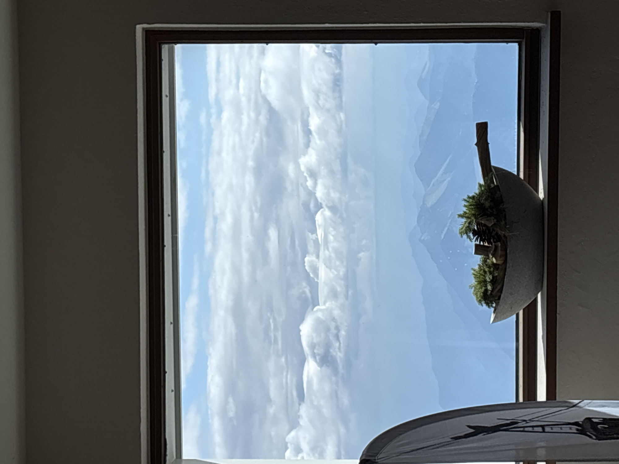 The image shows a view from a window in Ehrwald, Austria, overlooking a layered mountain landscape under a cloudy sky. In the foreground, on the windowsill, is a decorative bowl filled with greenery, pinecones, and a wooden stick. To the left of the bowl, partially visible, is a tall, white object with a black graphic, possibly a flag or banner. The window frame is brown, and the wall around it is a neutral, light brown color. The scene captures a serene and expansive view of the Austrian Alps.