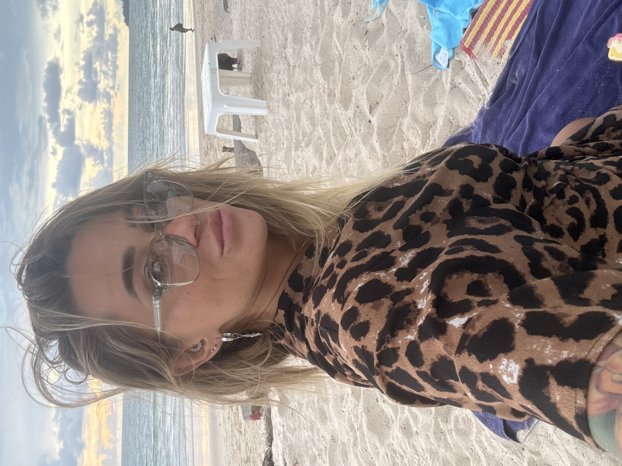 A woman with blonde hair and glasses is sitting on a beach in Choeng Thale, Thailand. She is wearing a leopard print top and is looking at the camera. The beach is sandy, and there is water in the background. There is also a white plastic table and chairs on the beach. The sky is cloudy with some sunset colors.