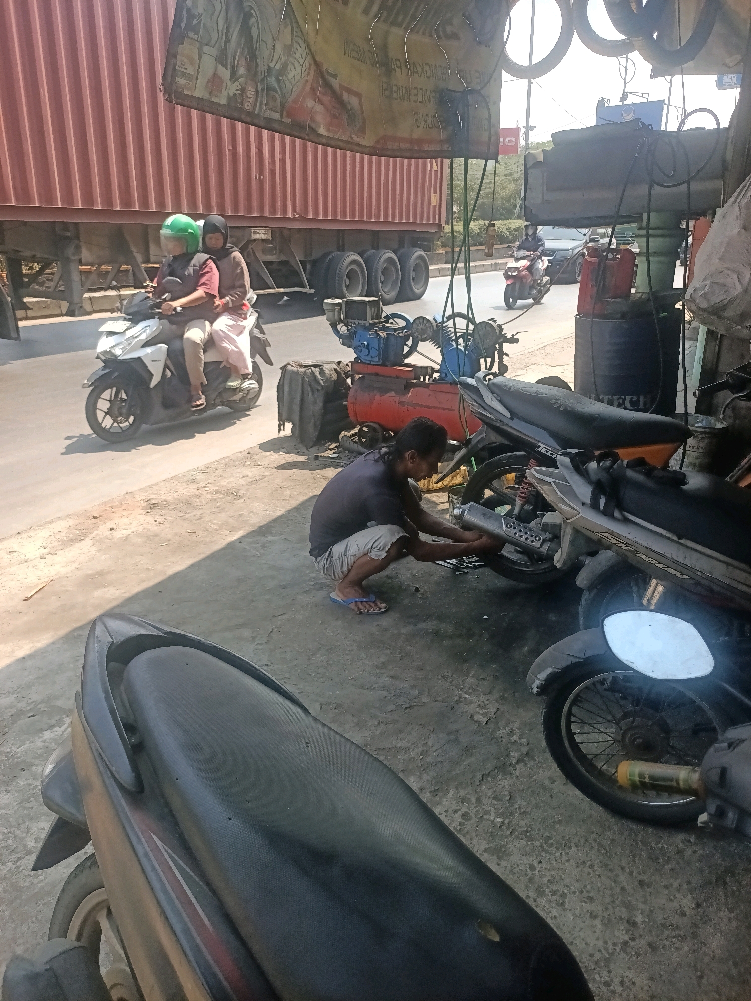The image depicts a street-side motorcycle repair shop in Gandasari, Indonesia. In the foreground, a mechanic, a man wearing a dark t-shirt and shorts, is crouched down, actively working on the exhaust pipe of a parked motorcycle. He appears to be using a tool to adjust or fix the pipe. To the left of the mechanic, another motorcycle is parked, partially visible, with its seat in the foreground.
In the background, a white scooter carrying two passengers is driving away on the paved road. The passenger in the front is wearing a green helmet and a dark vest over a maroon shirt, while the passenger behind is wearing a dark hijab and a light brown top. A large, red truck is parked or moving slowly on the same road, its trailer covered with a yellow tarp.
The environment appears to be an outdoor setting with a dusty, uneven ground surface. Various tools and equipment related to motorcycle repair, including what looks like an air compressor and drums, are visible in the vicinity of the shop. The overall scene suggests a bustling, working environment typical of a local repair business in Indonesia.
