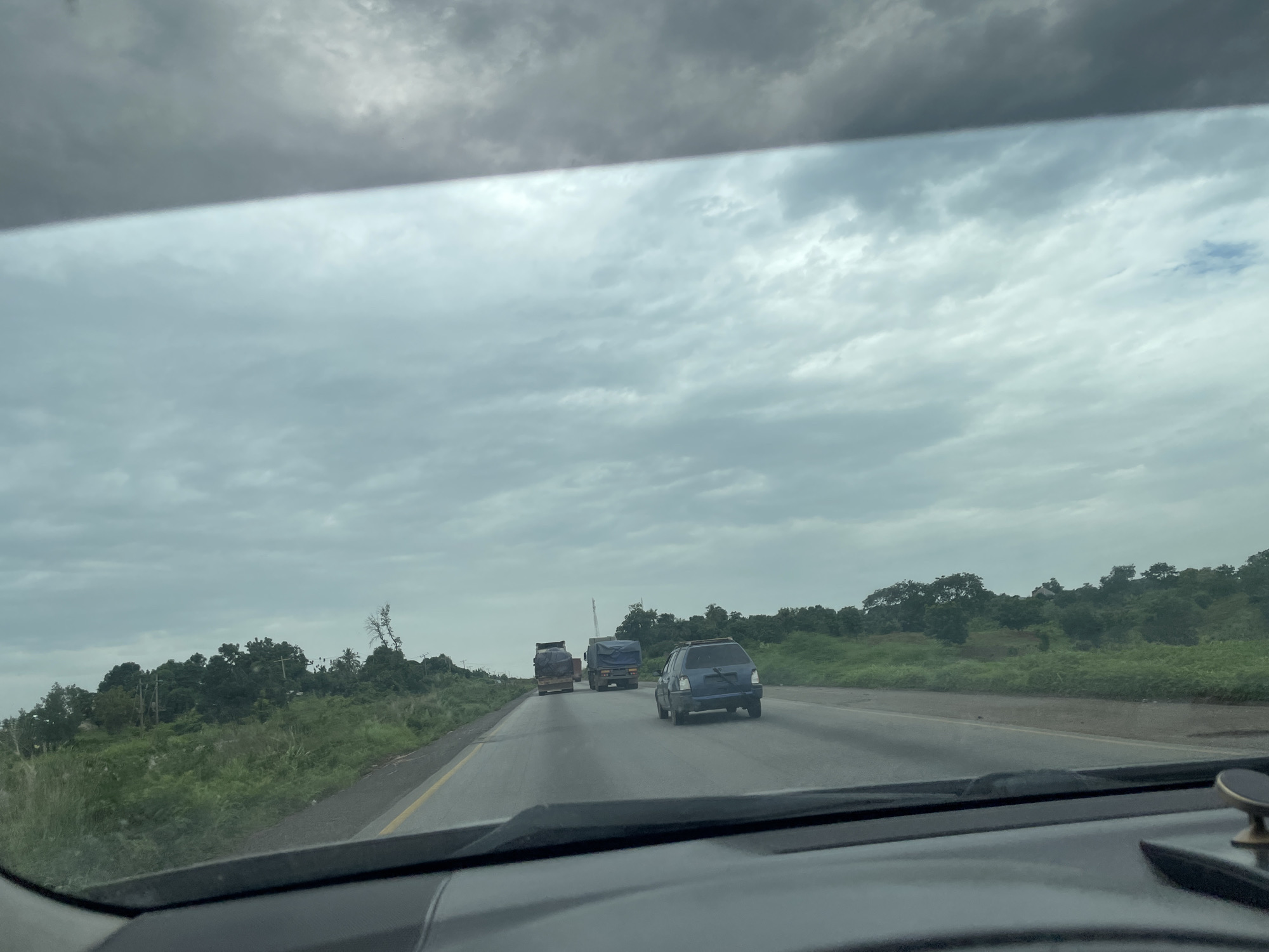 The image shows a view from inside a vehicle on a highway in Kwali, Nigeria. The sky is overcast with gray clouds. The highway is paved and has a yellow line marking the lanes. Several vehicles are on the road, including a blue hatchback and two trucks, one of which has a covered cargo. Lush green vegetation and trees line the sides of the highway, with some signs of human habitation in the distance, such as utility poles. The overall scene suggests a typical day of travel on a highway in a tropical region.