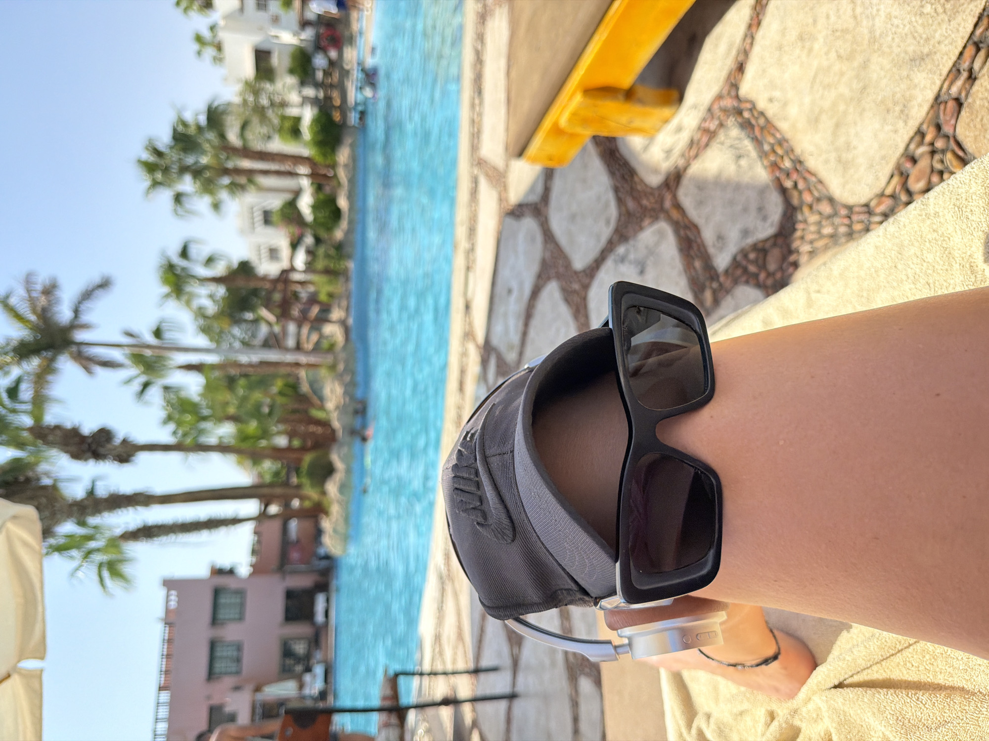 A close-up, first-person perspective shot shows a person's legs and lower face, partially obscured by a baseball cap and sunglasses. The person is lying on a beige towel on a lounger, with a large, blue swimming pool and palm trees in the background, suggesting a resort location in Sharm El Sheikh, Egypt. The ground around the pool is paved with large, light-colored stones interspersed with small, brown pebbles. In the distance, the pink facade of a building and white structures can be seen, typical of resort architecture in the region. The person is wearing silver headphones. Sunlight illuminates the scene, casting shadows and creating a bright, summery atmosphere.
