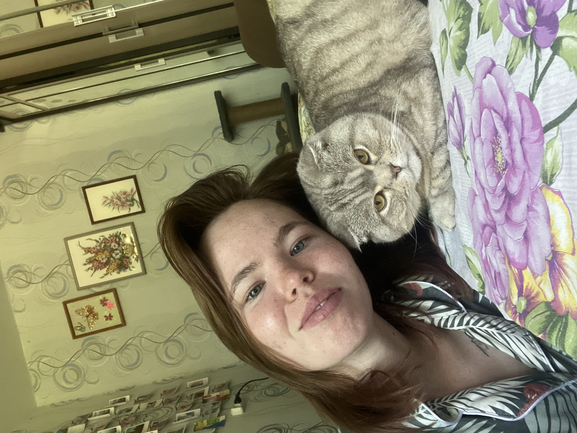 A young woman with auburn hair and freckles is taking a selfie with her Scottish Fold cat. They are lying on a bed with a floral-patterned comforter. The woman is wearing a black and white patterned pajama top. The cat is grey with tabby markings and has distinctive folded ears. In the background, there is wallpaper with a circular pattern, several framed pictures of flowers and a butterfly, and a tall, mirrored wardrobe. A cat scratching post is visible behind the cat. The room appears to be a bedroom. The scene is domestic and informal.