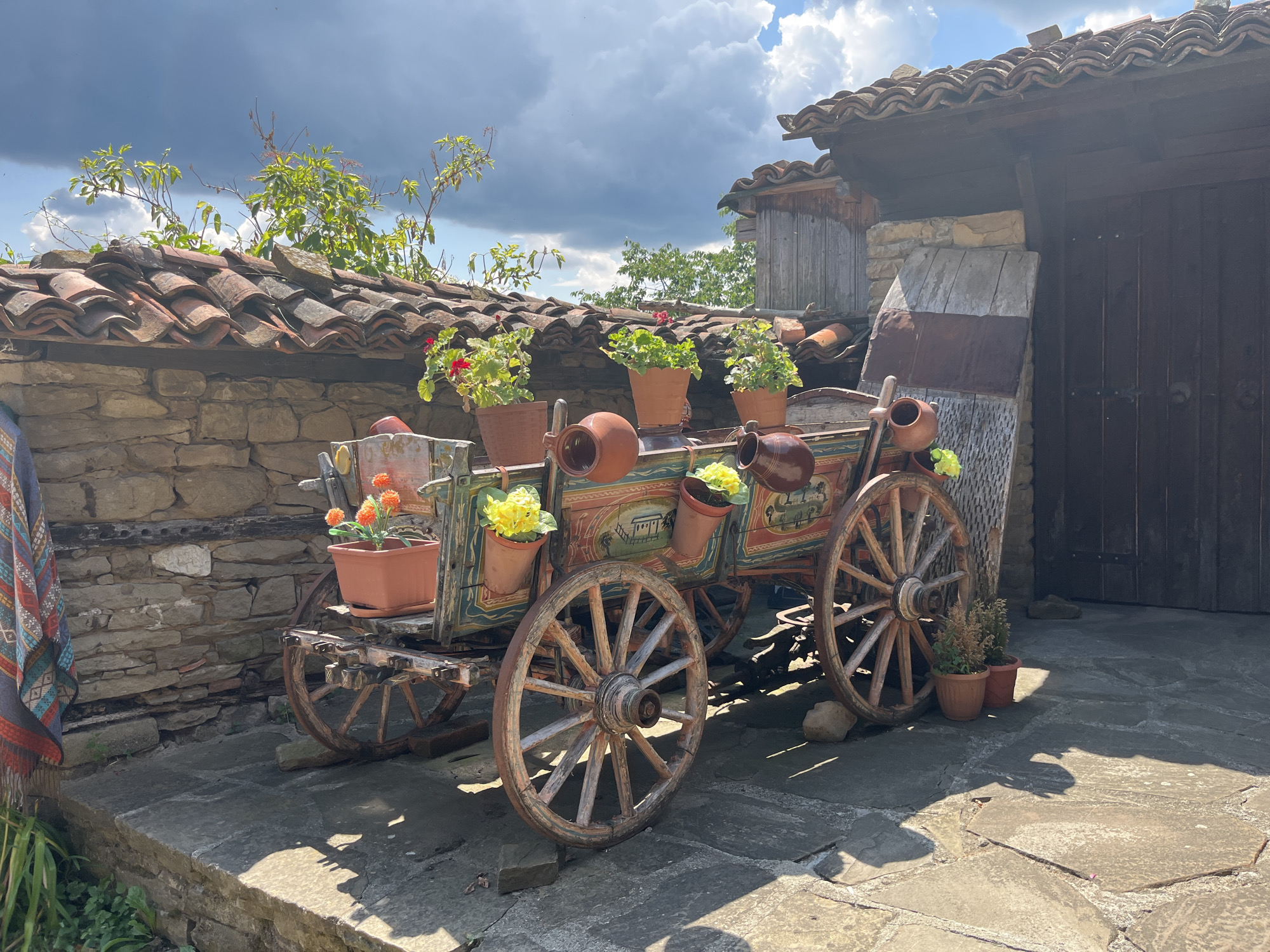 The image depicts an old, ornate, wooden wagon with painted designs, used as a decorative planter, situated on a cobblestone patio in Zheravna, Bulgaria. The wagon is adorned with several terracotta pots containing flowers, including red and yellow blooms. Behind the wagon is a stone wall topped with a weathered terracotta tile roof, characteristic of traditional Bulgarian architecture. A rustic wooden door is visible to the right of the wagon, adjacent to a stone and wood structure. The scene is bathed in sunlight, casting distinct shadows on the patio, and the sky above is partly cloudy. Green foliage and branches are seen peeking over the roofline, suggesting a natural, outdoor setting. The wagon appears to be a static decorative element, with no people or animals present.