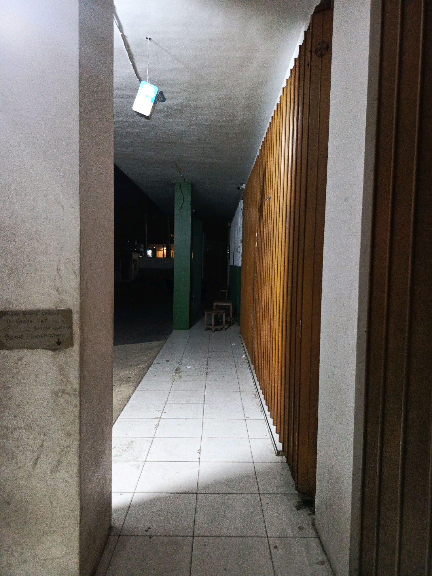 The image depicts an outdoor walkway in Pamekasan, Indonesia, at night. The walkway is paved with square tiles and is bordered by a light-colored concrete pillar on the left and a tall, wooden, slatted door on the right. A sign is affixed to the concrete pillar on the left, containing handwritten text in Indonesian, which appears to be a directive about not littering. Further down the walkway, two green pillars are visible. The ceiling above the walkway is rough concrete. A piece of what appears to be a blue plastic bag is suspended from the ceiling by a string. The area beyond the walkway is dark, with some faint lights visible in the distance, suggesting it is a street or a building entrance. Two wooden stools are placed against the green pillars. The scene is dimly lit, with shadows cast on the walkway from an unseen light source.