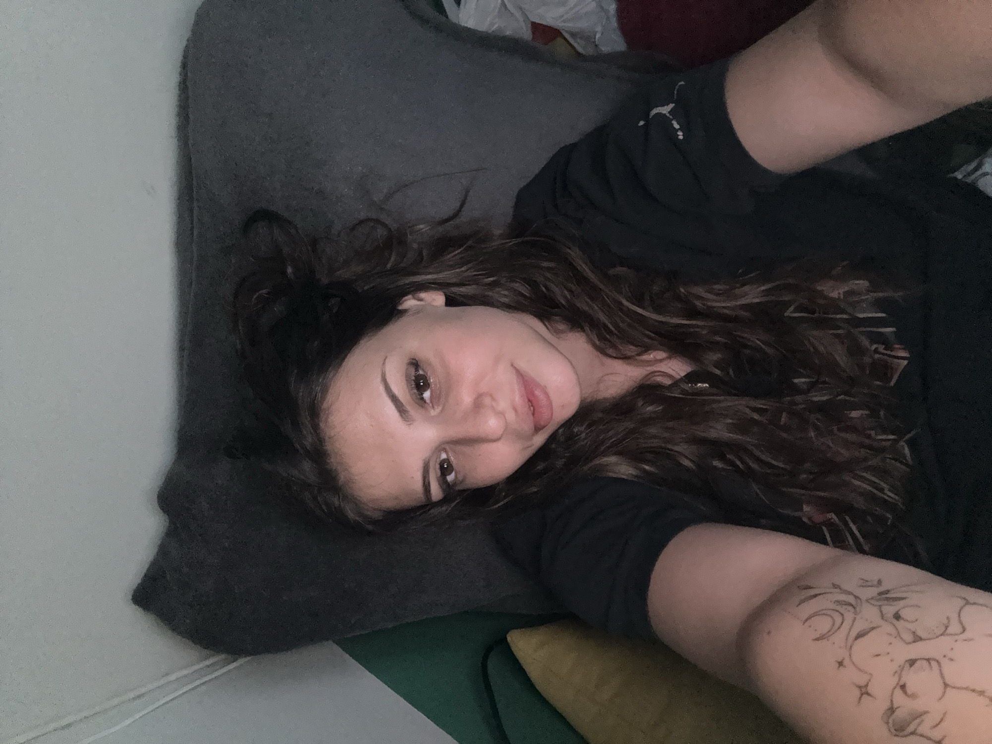 The image depicts a young adult female in an indoor setting. She is positioned as if lying down or reclining, looking directly at the camera with a slight smile, suggesting a selfie. Her long, dark, wavy hair is somewhat disheveled around her face and shoulders. She has brown eyes and full lips, with visible eyeliner and mascara. She is wearing a short-sleeved black top, with a small white logo resembling a stylized athletic figure visible on her right sleeve near the shoulder. A detailed outline tattoo is present on her left upper arm, featuring two felines (possibly lions or similar big cats) facing each other, surrounded by celestial elements including a crescent moon, stars, and abstract foliage. The background consists of a dark grey upholstered surface, likely a pillow or sofa back, behind her head and to her right. To her left, a green upholstered surface is visible, partially obscured by a yellow cushion or pillow. Further to her right, beyond her arm and the dark grey surface, indistinct items, some appearing white or light-colored, are visible against a darker background. Above her head is a light-colored wall or ceiling, with a thin white cord running vertically along the left edge. The lighting appears somewhat dim. The image depicts an indoor scene; however, no visual information is present to identify the specific location as Plovdiv, Bulgaria.