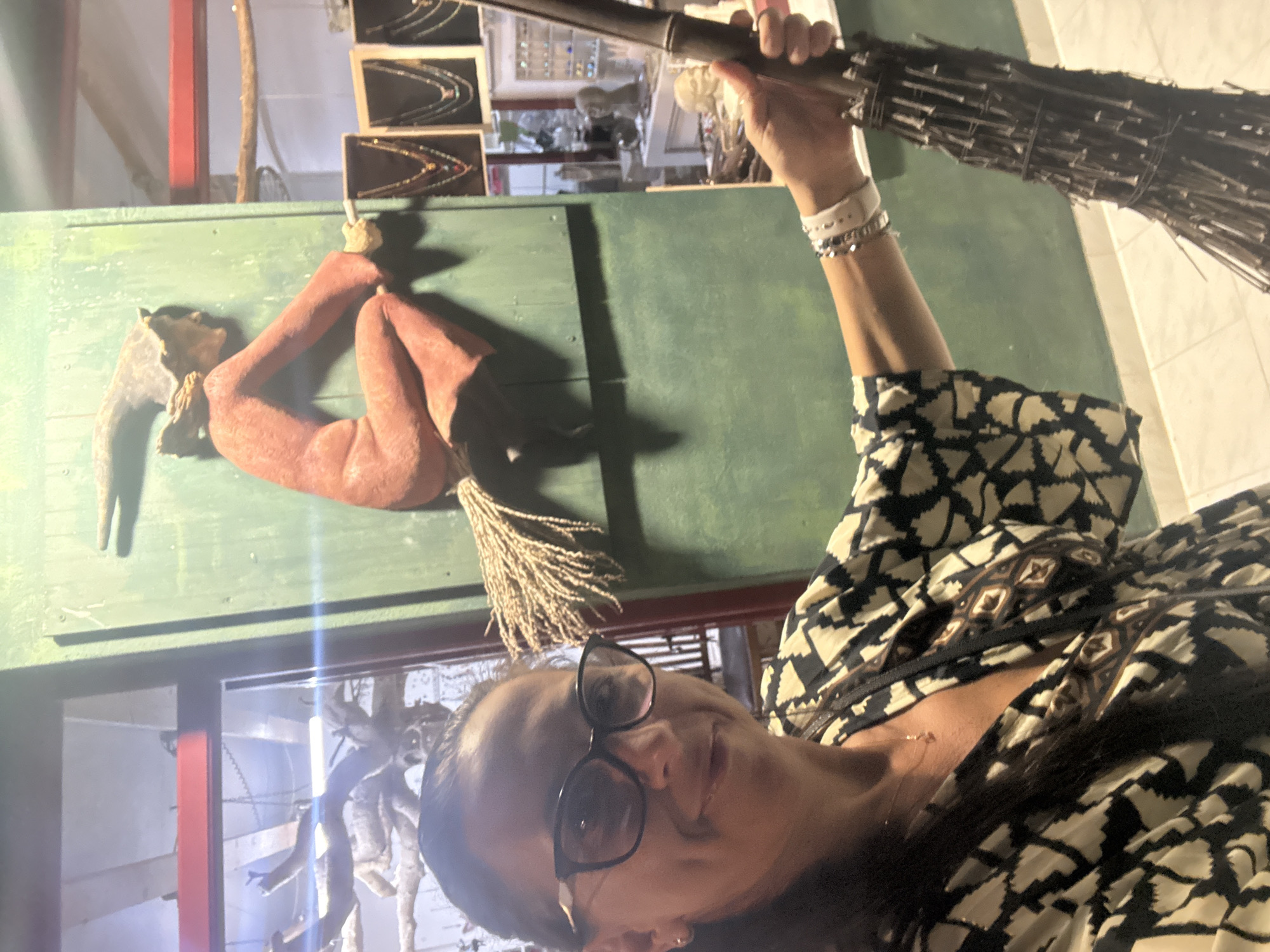 A woman wearing glasses and a black and white patterned dress stands in front of a green wall. She is holding a broomstick in her right hand. Behind her, a figurine of a witch on a broomstick is mounted on the wall. To the right of the witch figurine, several necklaces are displayed on stands. The background also shows a display of busts and other decorative items. The flooring appears to be tiled. The scene is indoors, likely a shop or a themed exhibit. The location is identified as the city of Pelekas, Greece.