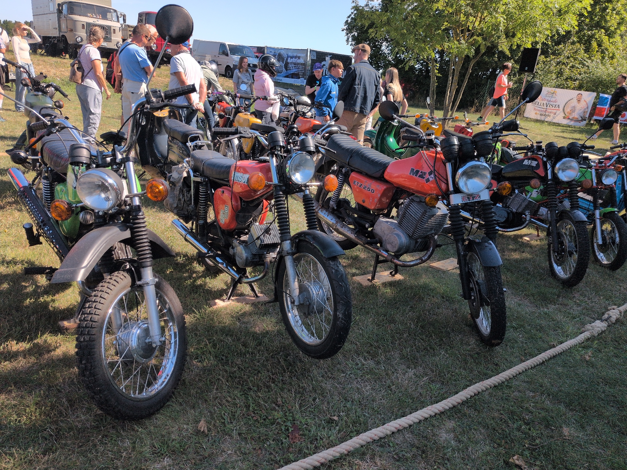 This image captures an outdoor event, likely a motorcycle exhibition or gathering, in Hohenferchesar, Germany. The foreground is dominated by several vintage motorcycles parked on a grassy area. The motorcycles are a mix of brands and colors, with many featuring prominent chrome details and retro designs. One motorcycle in the center is orange with the designation "MZ ETZ 250" visible on its side. Another in the foreground is predominantly green.
In the background, a gathering of people is observed, milling around the motorcycles and interacting with each other. They are dressed casually in t-shirts, jeans, and some are wearing hats. Some individuals appear to be examining the motorcycles, while others are engaged in conversation. Behind the crowd, various vehicles are visible, including a large truck, vans, and what appears to be part of a banner or advertisement. The environment is sunny with clear blue skies, suggesting fair weather for the event. The presence of trees in the background indicates a park or open field setting. A thick rope lies coiled on the grass in the lower right corner of the frame, possibly demarcating an area or for a specific activity.