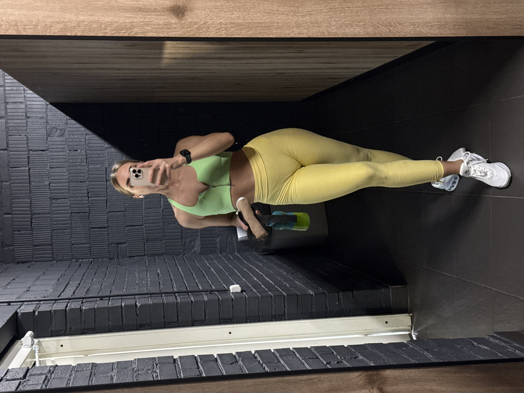 A woman in a lime green sports bra and yellow leggings is taking a selfie in what appears to be a gym or workout space. She is holding her phone in her left hand, and it obscures part of her face. She is wearing a smartwatch on her left wrist. She is standing in front of a black brick wall, with a white window frame below. A wooden ceiling panel is visible above. Her right foot is forward, and she is wearing white sneakers. Behind her, a black trash can is visible, with a cleaning spray bottle resting on it. The floor is dark grey. There is no indication of the city or country.