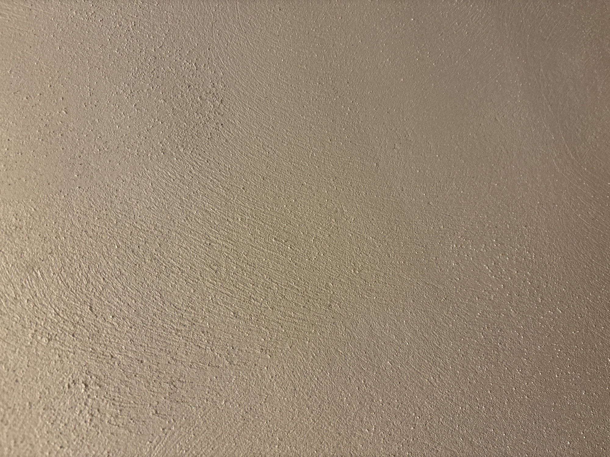 The image displays a close-up view of a textured beige wall. The texture consists of small, raised bumps and subtle, sweeping lines, suggesting a stucco or rough plaster finish. The lighting appears to be soft and casts minimal shadows, highlighting the uneven surface. There are no people, objects, or discernible environmental features that would allow for the identification of a specific location within Burgas, Bulgaria.