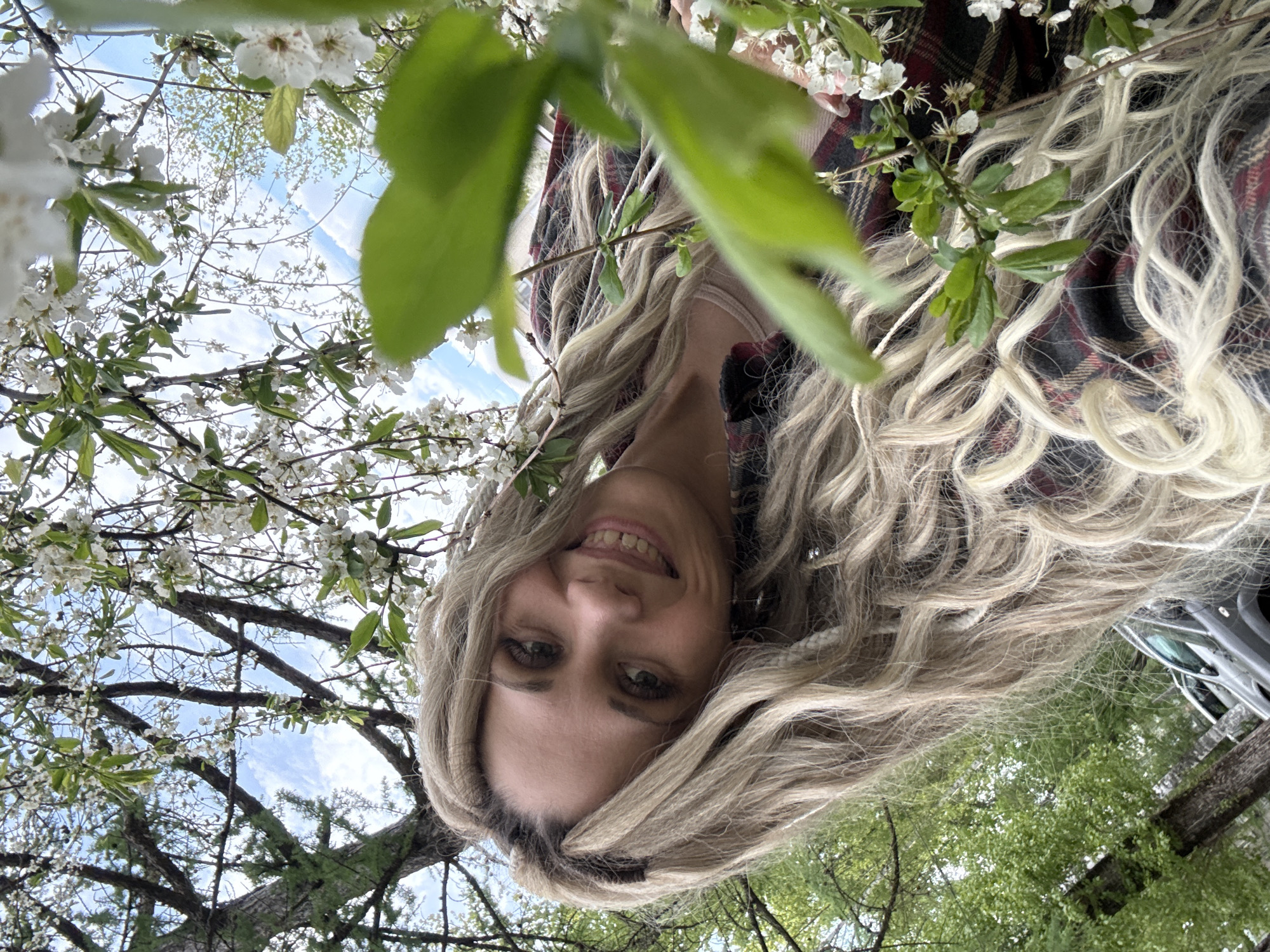 A woman with long, blonde, wavy hair is smiling at the camera. She is wearing a red and black plaid shirt. The shot is taken from a low angle, looking up at her face, which is partially framed by branches of a tree with white blossoms. The sky is visible through the branches. Part of a car is visible on the right side of the image, at the top. The location is potentially in a park or garden setting based on the visible trees and foliage. The location of Rusanovo, Russia can't be confirmed from the content of the image.
