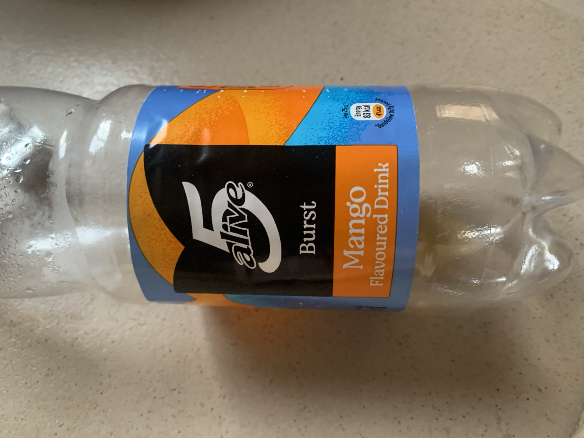 A partially empty, clear plastic bottle of 5 Alive Burst Mango Flavoured Drink is positioned vertically on a light-colored, textured surface. The bottle has condensation droplets on its exterior. The label is primarily black with white text, featuring the "5 alive" logo prominently. Below this, "Burst" is written, followed by "Mango Flavoured Drink" in white text on an orange band. A small section on the right of the label shows nutritional information, indicating "Energy 83 kcal" and "4% GDA" within a blue area. The background is a mottled, beige-toned wall. The lighting is neutral, casting soft shadows. No people or animals are present. The location is unknown.