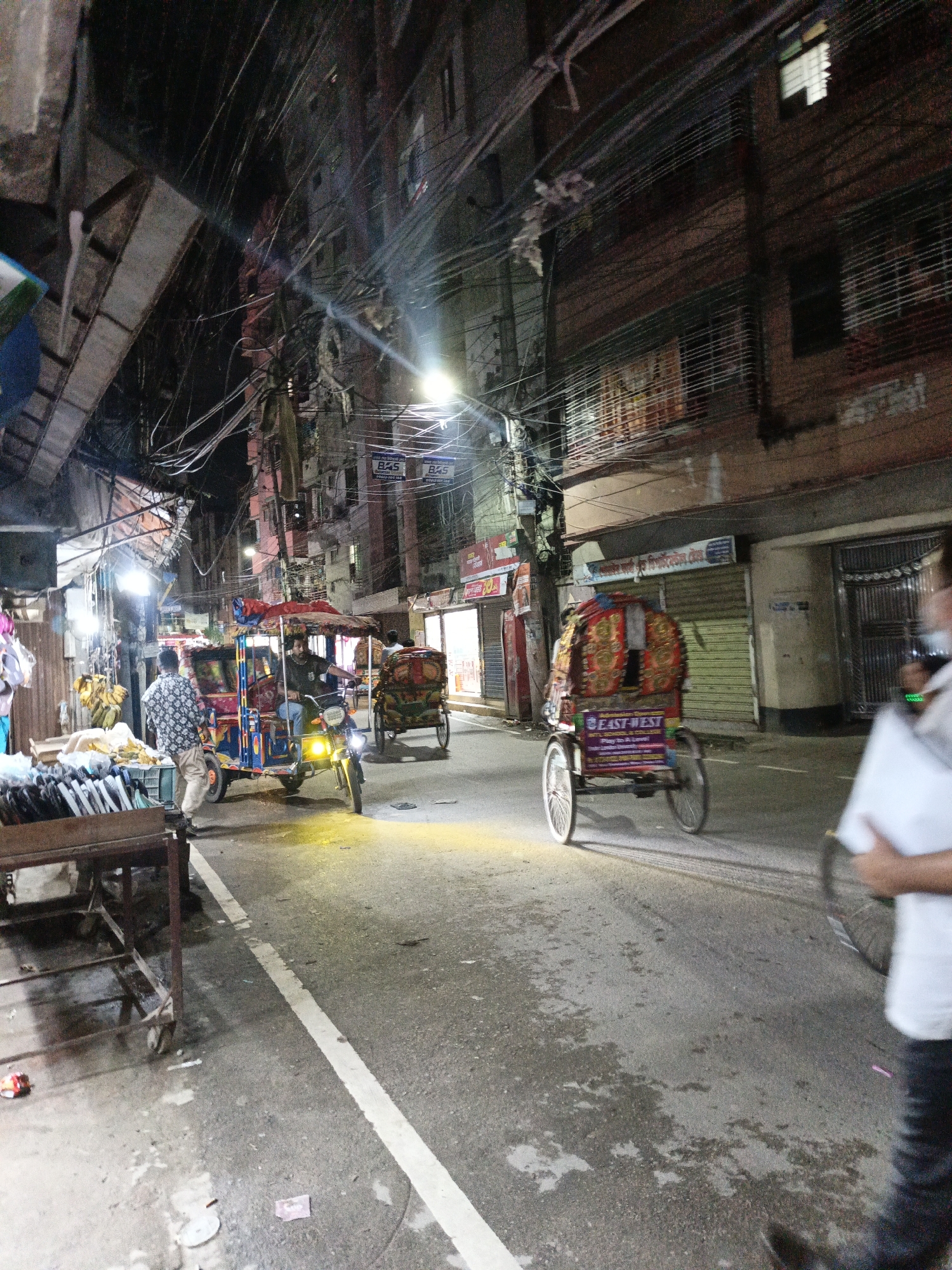 The image captures a street scene in Dhaka, Bangladesh, likely at night, given the artificial lighting and shadows. The street is paved and marked with a white line. Several vehicles are present, including two cycle rickshaws, one of which is brightly decorated with red and gold fabric and has the text "EAST-WEST" prominently displayed on its side. Another, smaller rickshaw, appears to be a motorized variant, also adorned with colorful decorations and lit by its headlights.
On the left side of the street, there is a vendor's stall with goods displayed, possibly food items like bananas and other produce. A man in a patterned shirt and light-colored pants stands near the motorized rickshaw, seemingly interacting with its driver. Another person, partially visible, is seated on the motorized rickshaw. In the background, buildings line the street, exhibiting a dense urban environment with numerous wires and cables suspended overhead. The buildings have windows and shutters, some of which are illuminated. A figure, out of focus in the foreground on the right, appears to be walking away from the camera, holding a white object. The overall atmosphere suggests a busy urban market or thoroughfare at dusk or night.