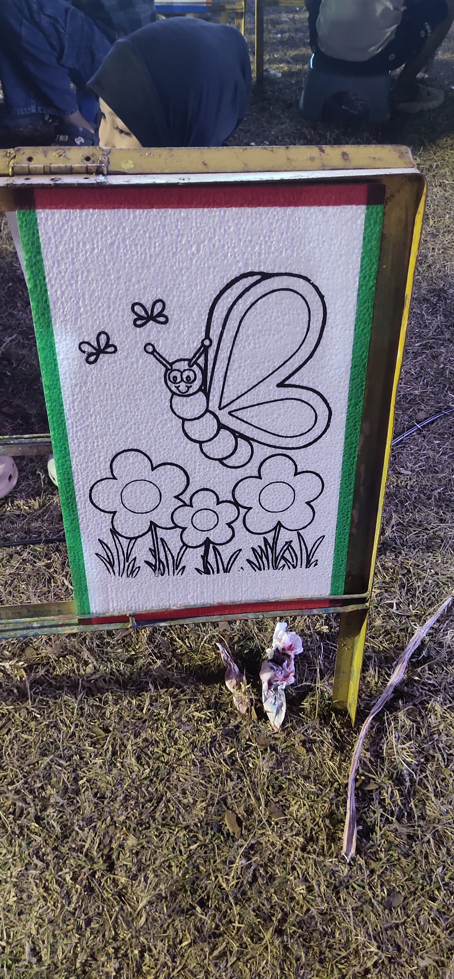 The image captures a close-up view of a white board with a black outline drawing of a butterfly and three flowers, positioned on a grassy outdoor surface. The board is framed by green and red tape on its left and top edges, respectively, and is supported by a yellow metal frame. In the background, blurred figures are visible, including a person wearing a dark headscarf and blue jeans. The ground is covered in dry grass and some scattered debris, including what appears to be wilting flowers or crumpled paper. The scene suggests a public space, possibly a park or an event ground, given the presence of the drawing board and the casual attire of the people in the background. The lighting indicates it might be evening or a dimly lit area. There is no specific indication of the city of Tasikmadu, Indonesia, within the image itself; this information is provided contextually.