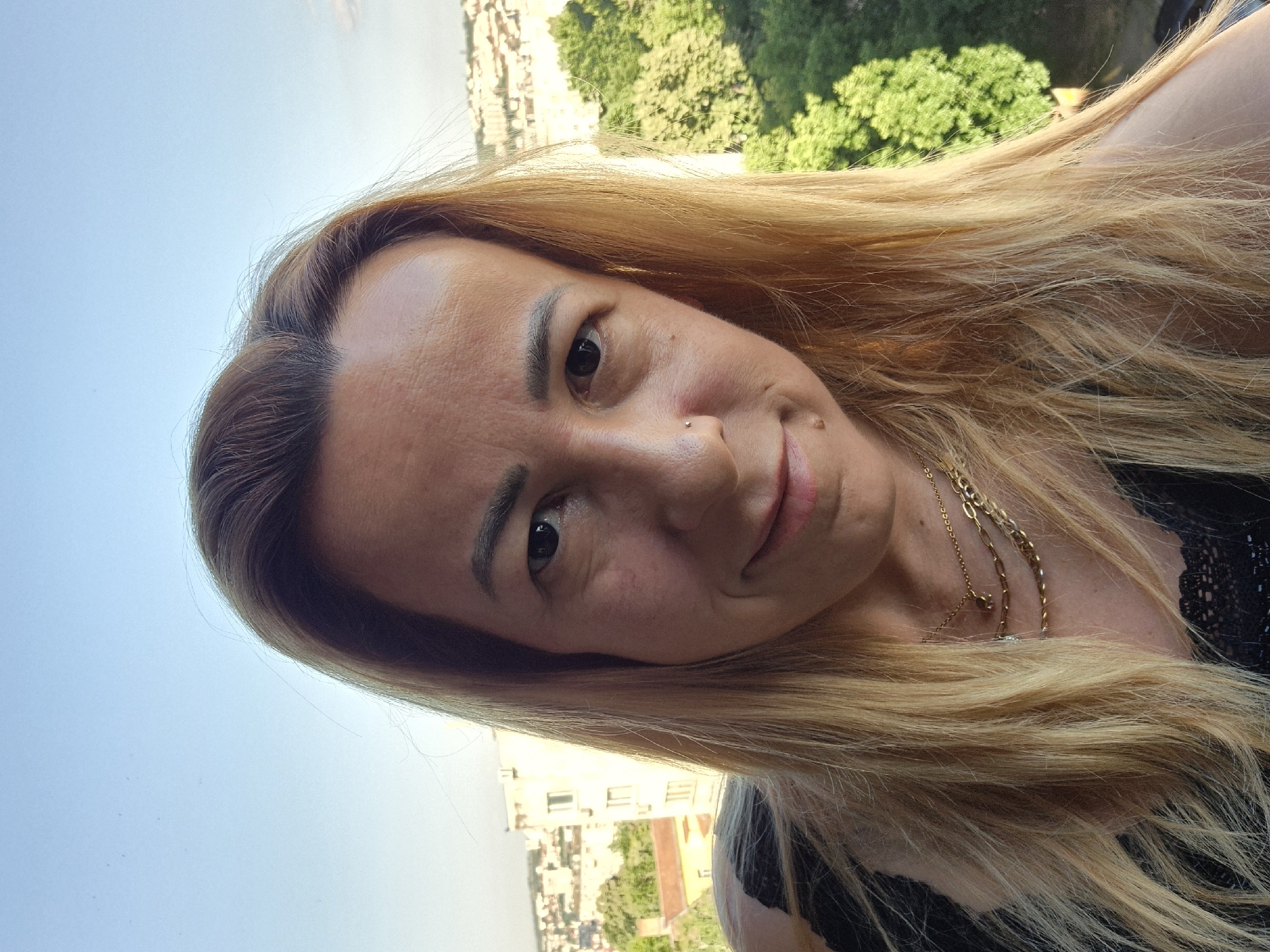 A woman with long blonde hair is taking a selfie outdoors with a city skyline and trees in the background. She is wearing a black top with lace detailing and several gold necklaces. The woman has a nose ring and is smiling at the camera. The setting appears to be a balcony or elevated position with buildings and lush green trees visible. The sky is a clear blue, suggesting it is daytime.