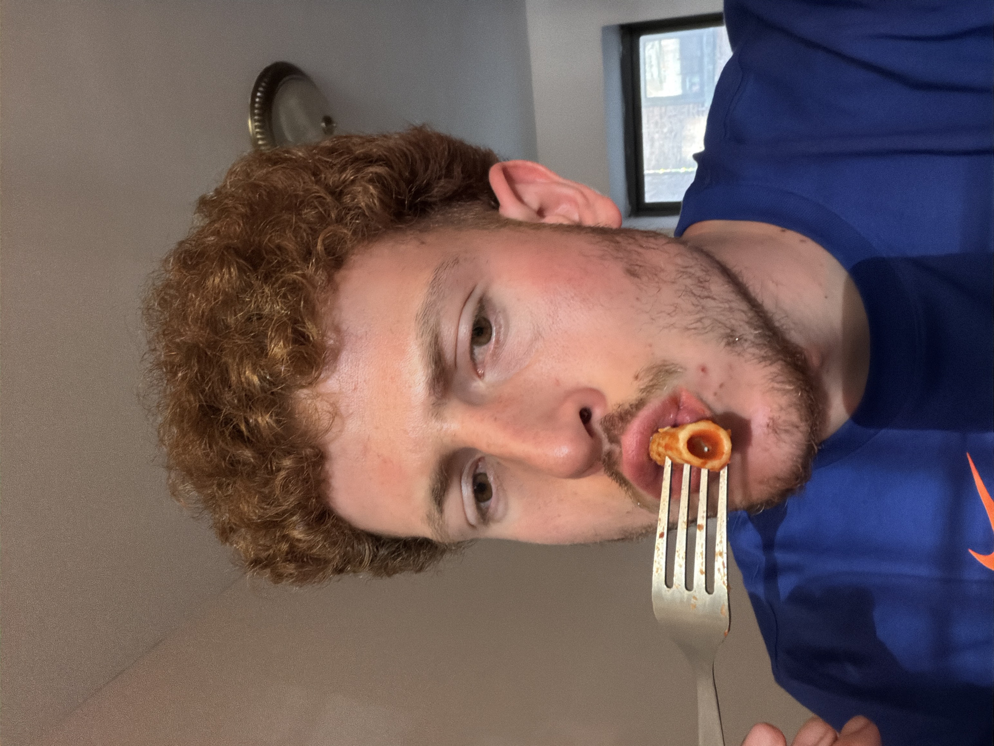 A fair-skinned man with curly red hair and a beard is eating a piece of pasta off a fork. He is wearing a blue Nike t-shirt. Behind him is a wall with a round wall-mounted light fixture and a window. It is not possible to determine if the location is in New York City.